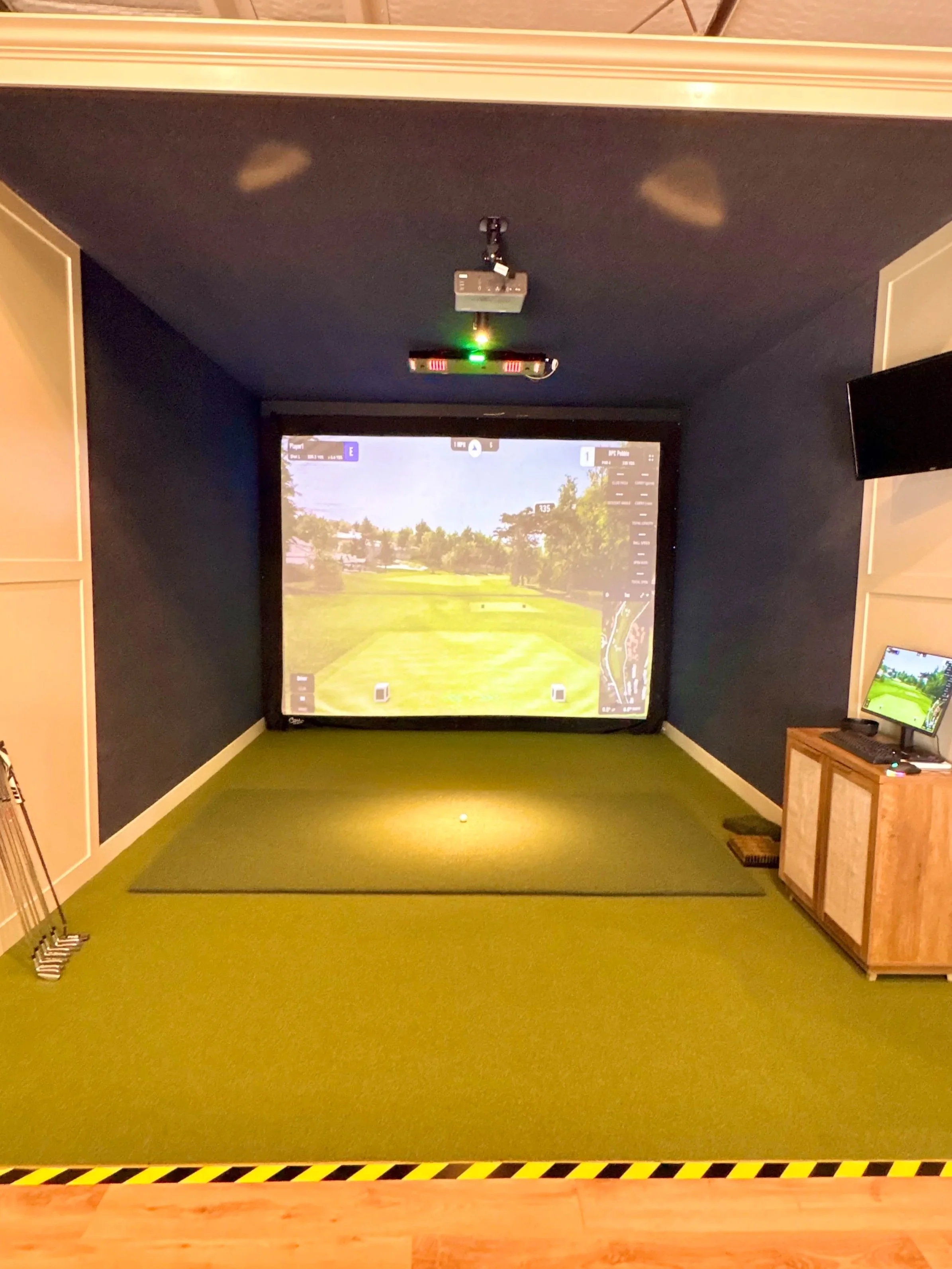 Indoor golf simulator with a screen displaying a golf course and a golf club and bag on the left side