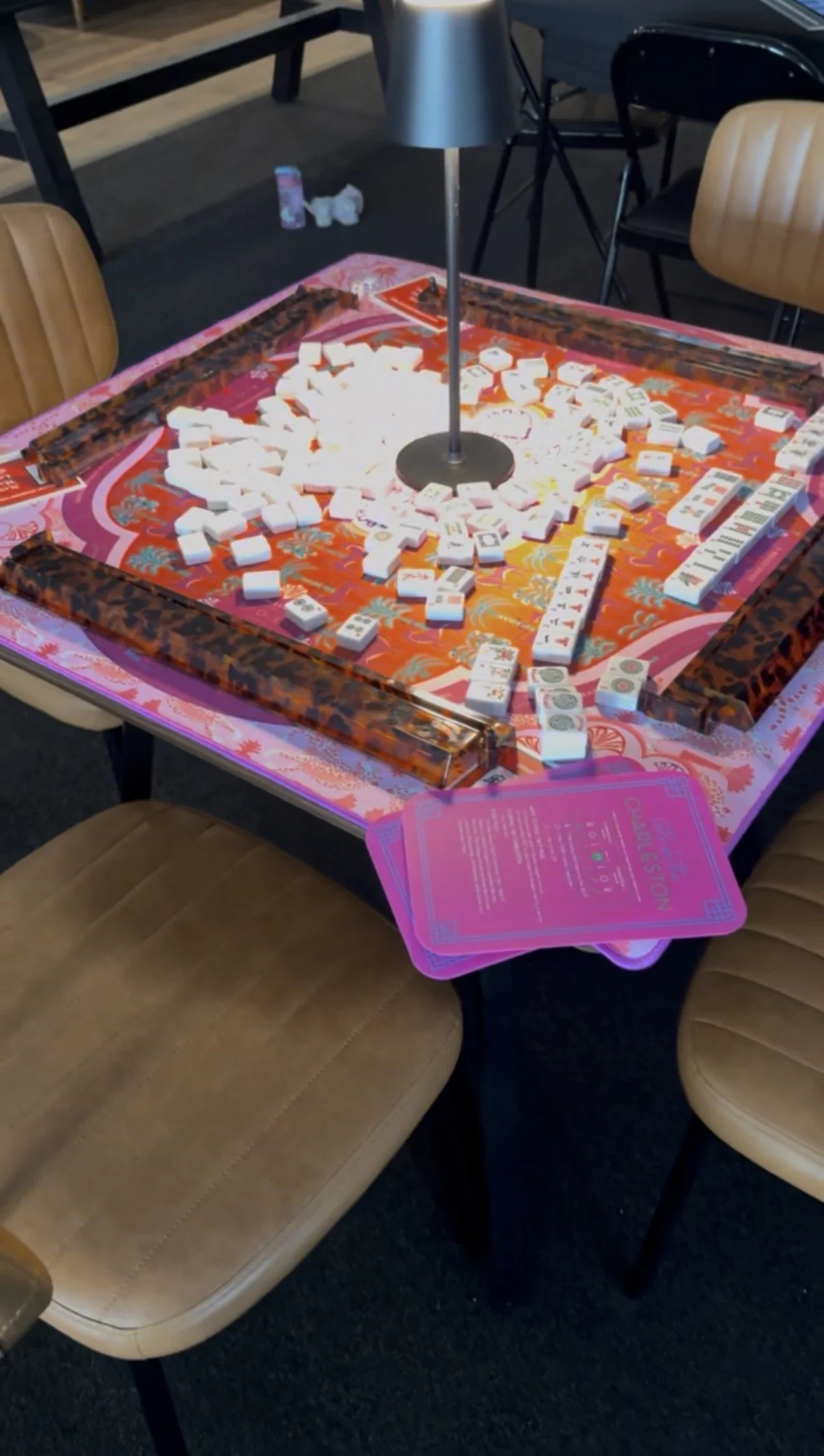 A table with scattered domino tiles, some with colorful markings and others with uniform white surfaces. There is a pink playing surface with a decorative border, a floor lamp, and chairs surrounding the table.