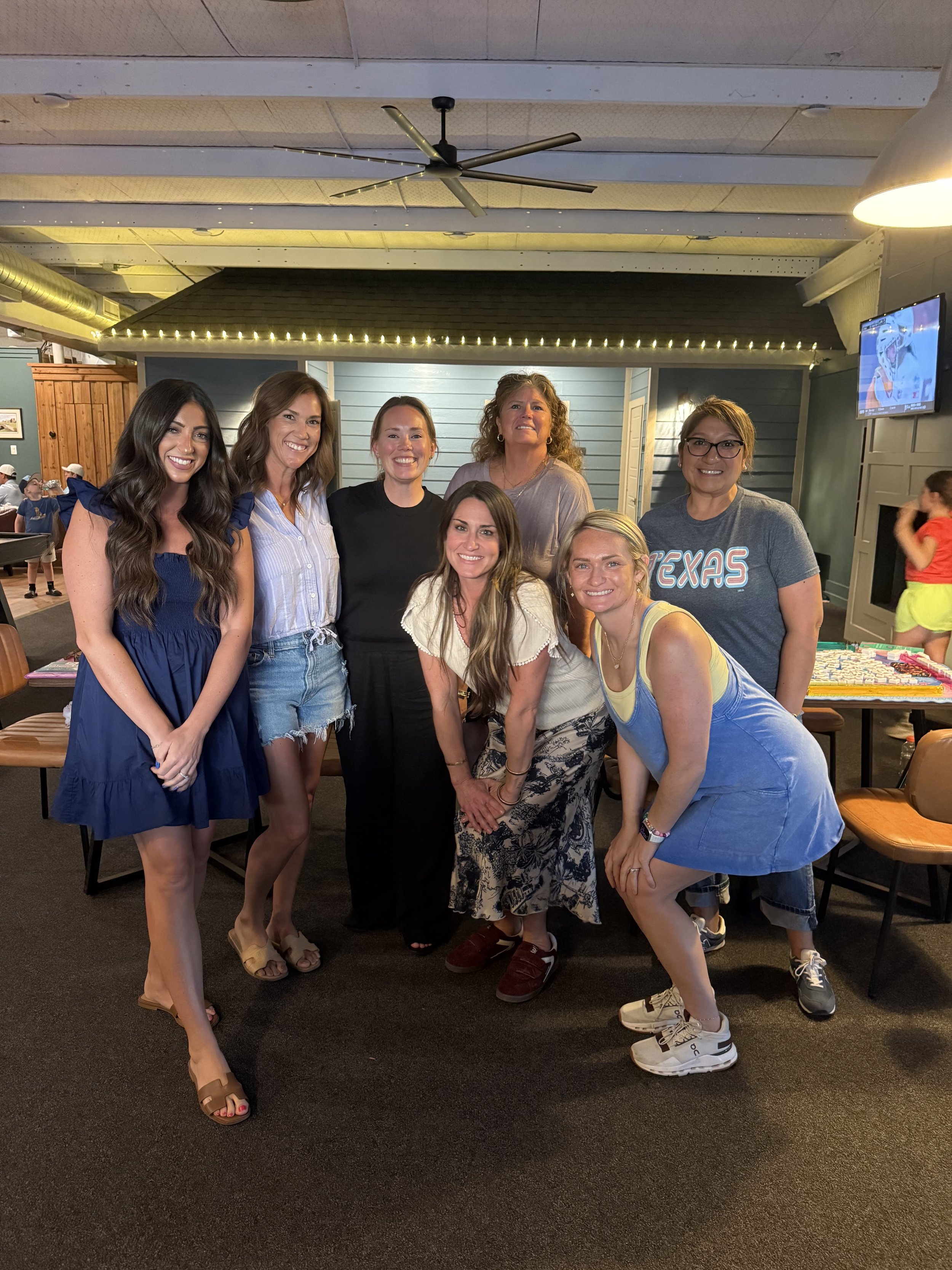 A group of six women standing close together in a restaurant or cafe, smiling at the camera. They are dressed in casual summer clothing, with some wearing dresses and others in shorts and tops. The background features a small building-like decor with string lights and a television on the wall displaying a cartoon.
