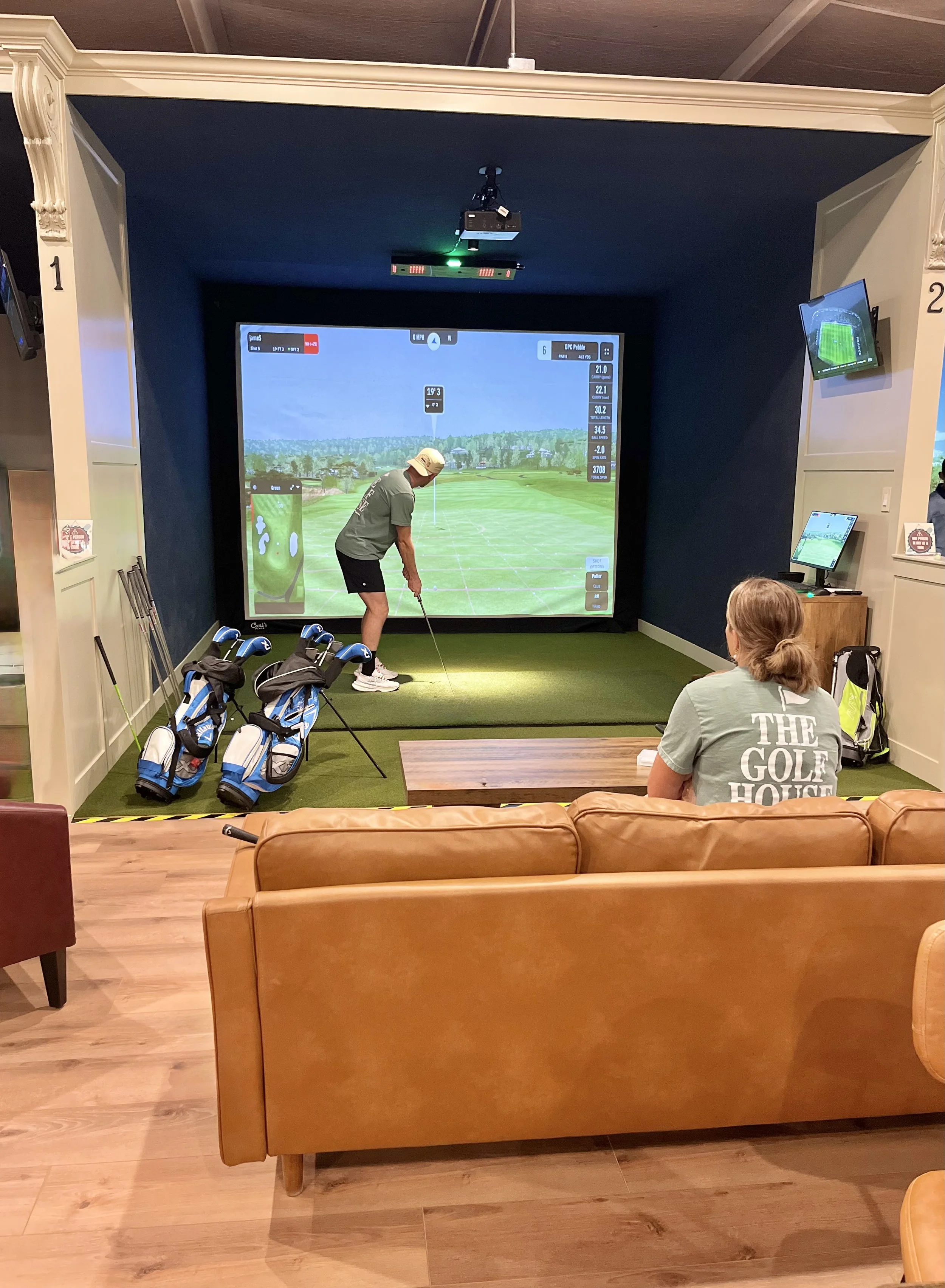 Person in pink shirt and yellow shoes practicing golf indoors on a green mat, preparing to putt.