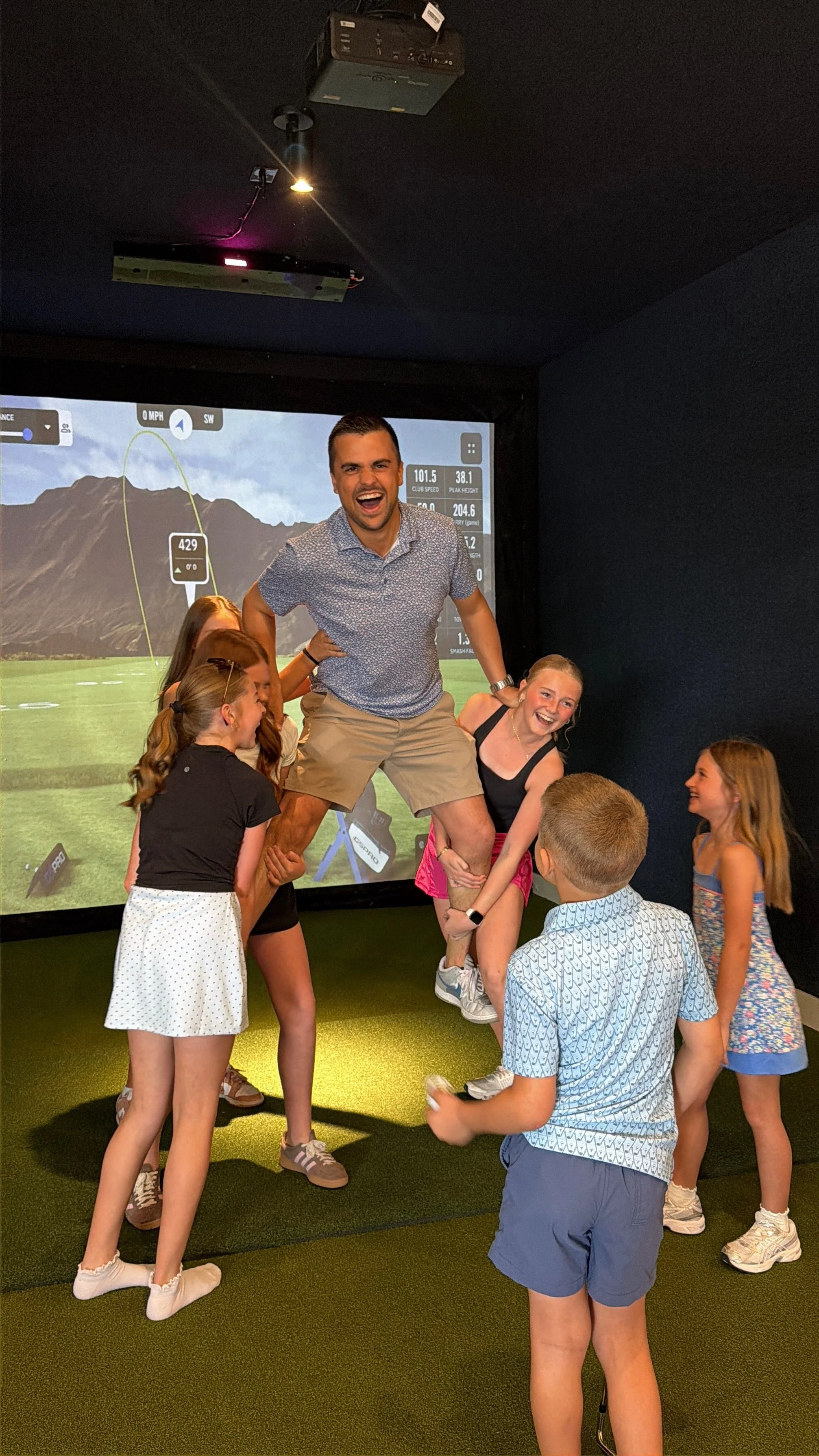 A group of children and two adults enjoying a game of indoor golf in front of a large screen displaying a golf course.
