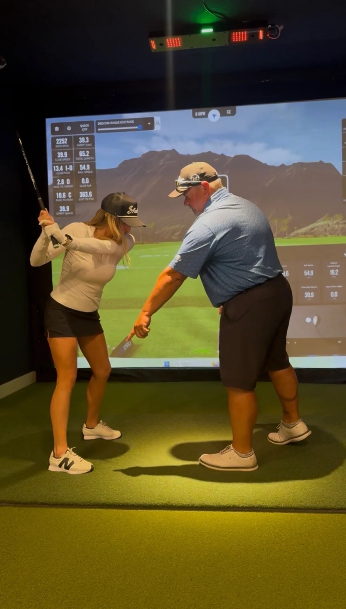 A man and a woman are playing indoor golf in front of a screen displaying a golf course scene with mountains, with data and stats on the sides. The man is wearing a blue shirt, black shorts, and a cap, while the woman is dressed in a beige long-sleeve shirt, black shorts, and white sneakers, holding a golf club.