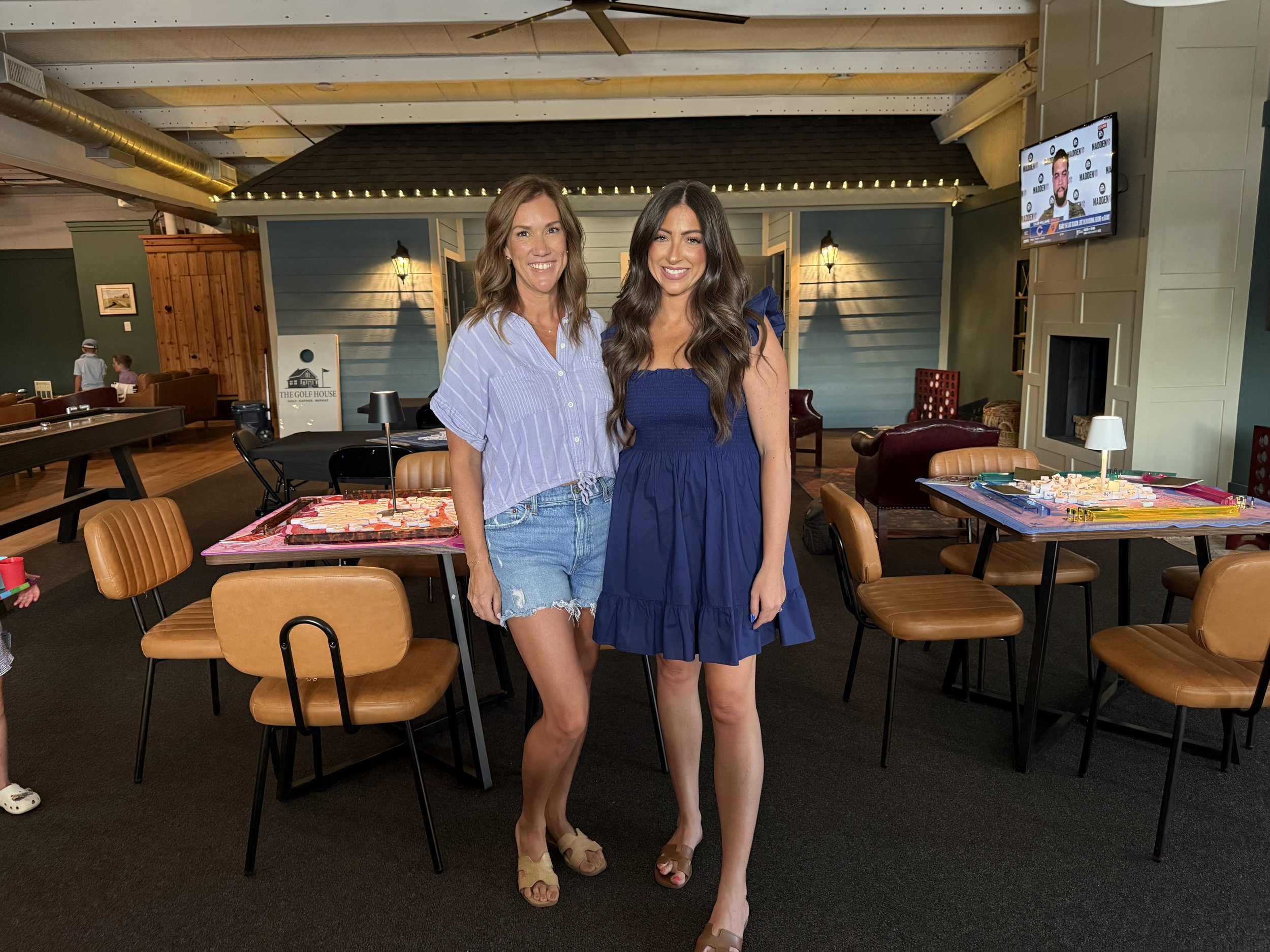 Two women standing inside a restaurant with tables and chairs, smiling at the camera. One woman has long brown hair and is wearing a blue dress, the other has shoulder-length light brown hair and is wearing a striped blue shirt and ripped denim shorts.