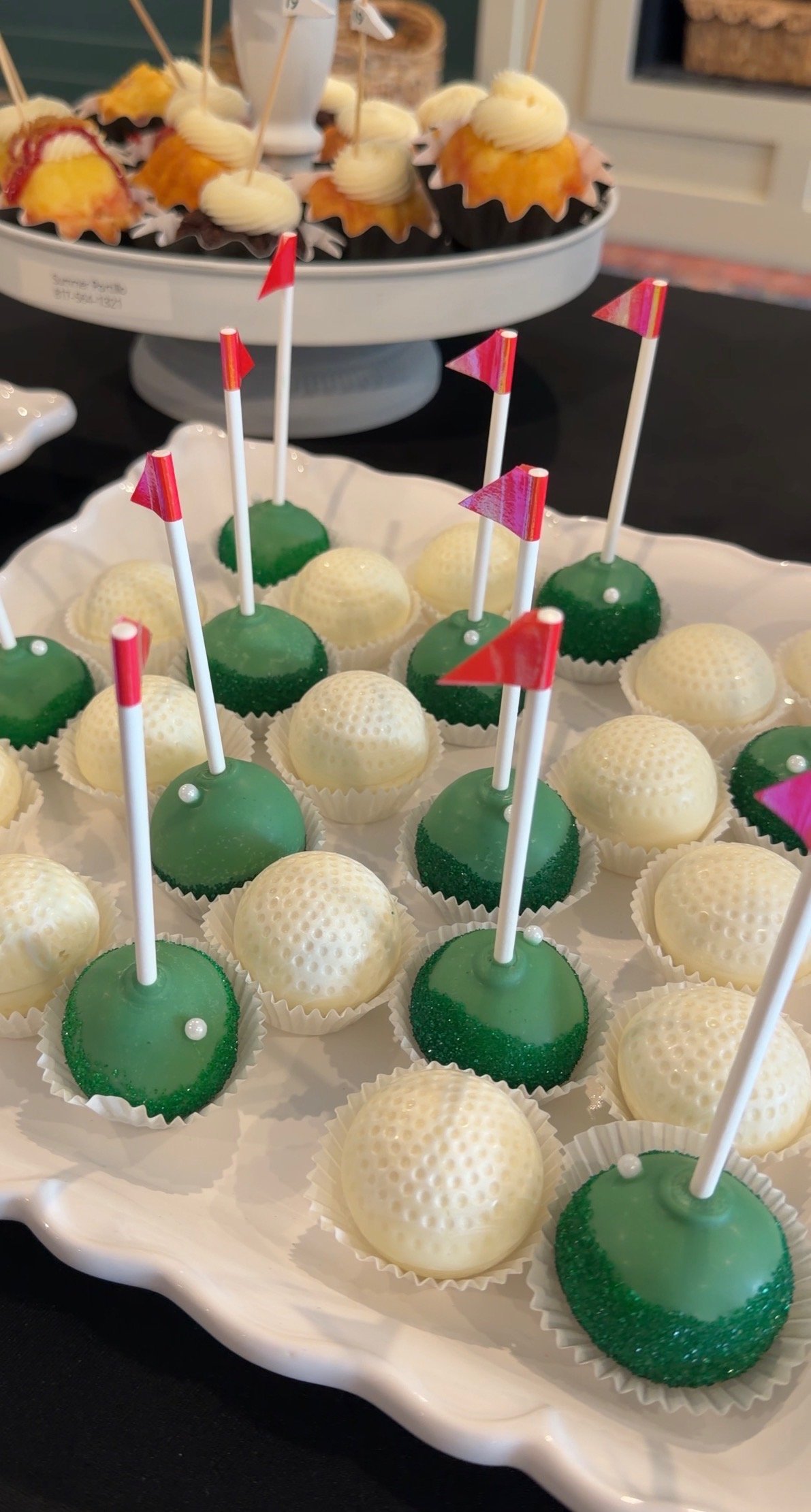 Tray of golf ball-themed cake pops with white and green coatings, each topped with a small flag, and a separate stand with cupcakes decorated with golf ball frosting and cream swirl toppers.