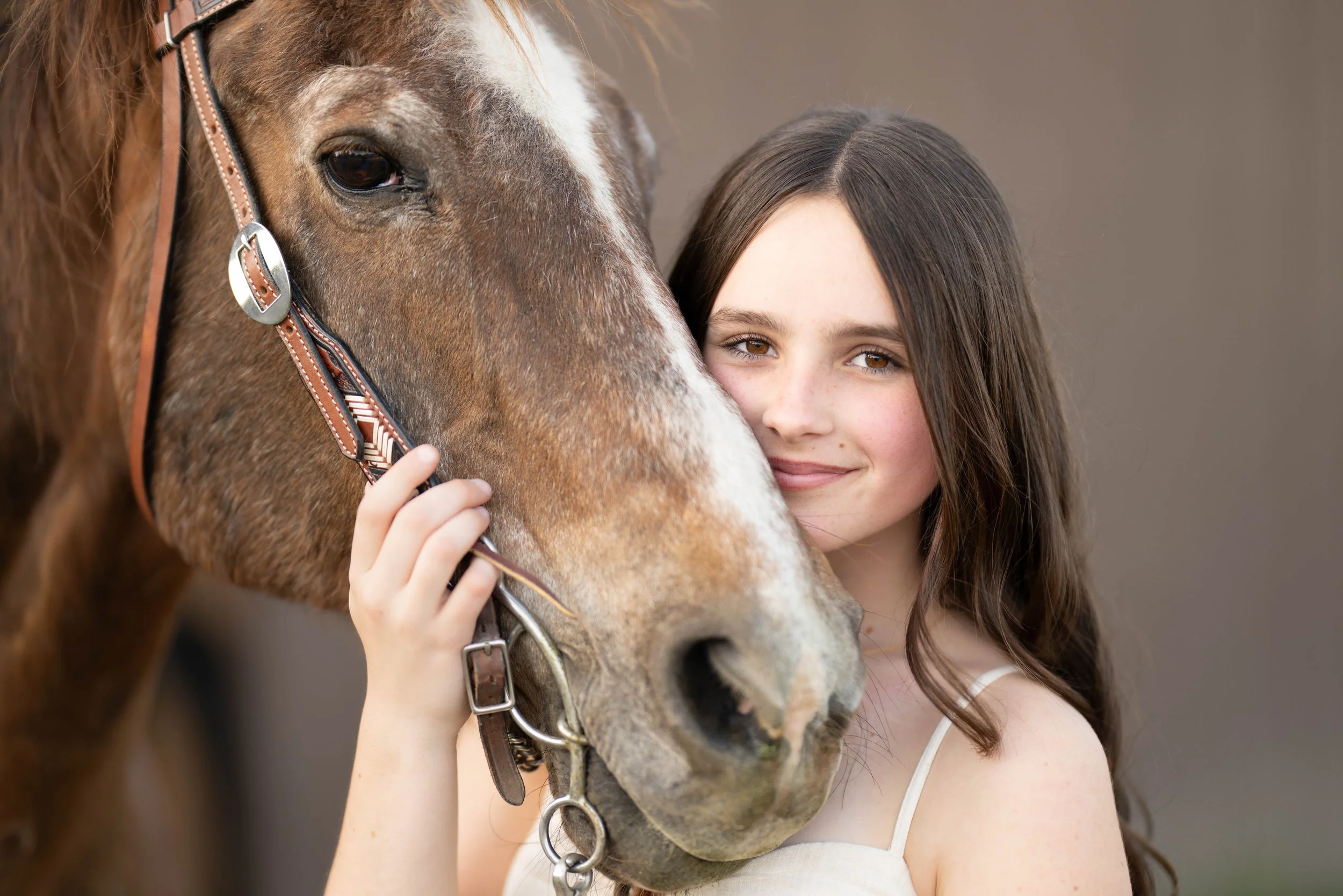 A young girl with long, dark hair hugging a brown and white horse, smiling softly, with a neutral background.