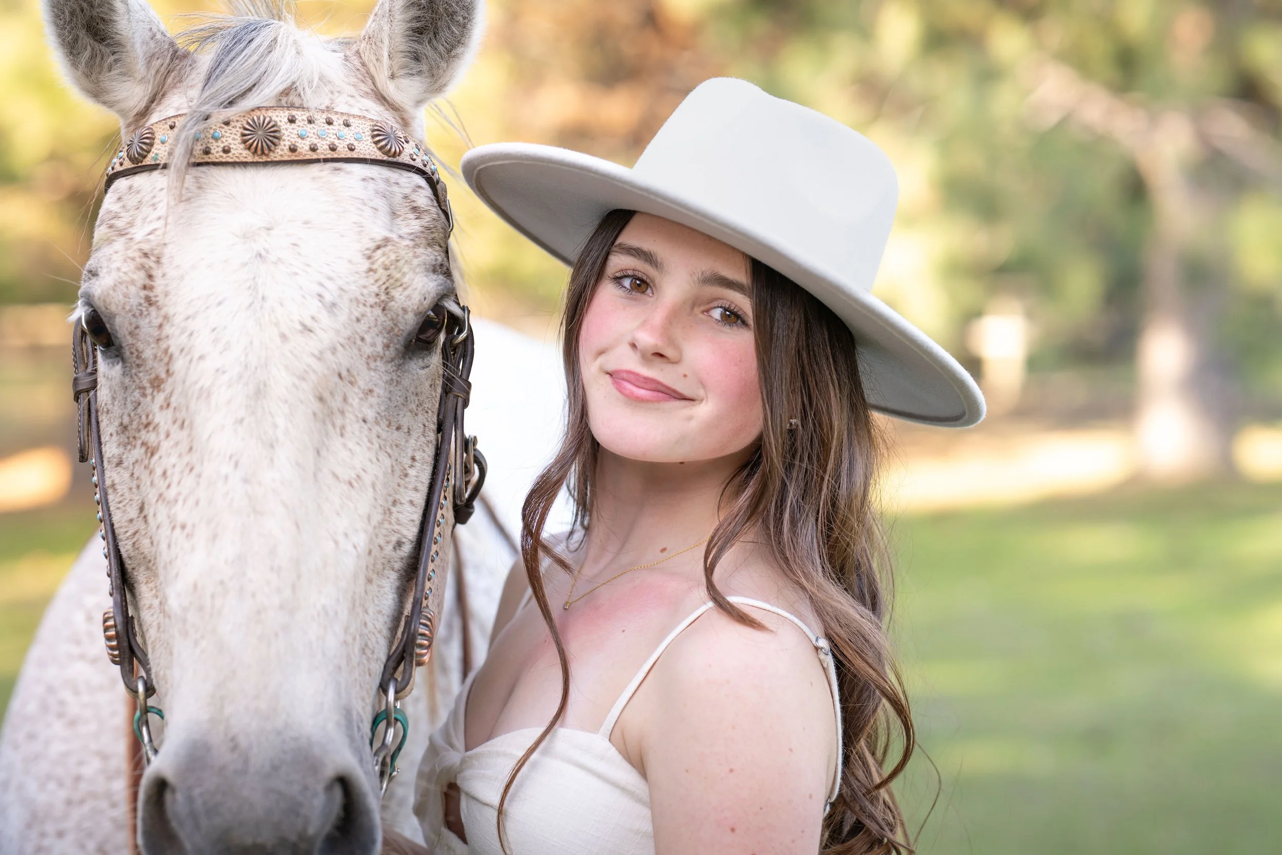 A young woman in a white dress and a wide-brimmed white hat standing next to a white horse with a decorated bridle in an outdoor setting with blurred green trees in the background.