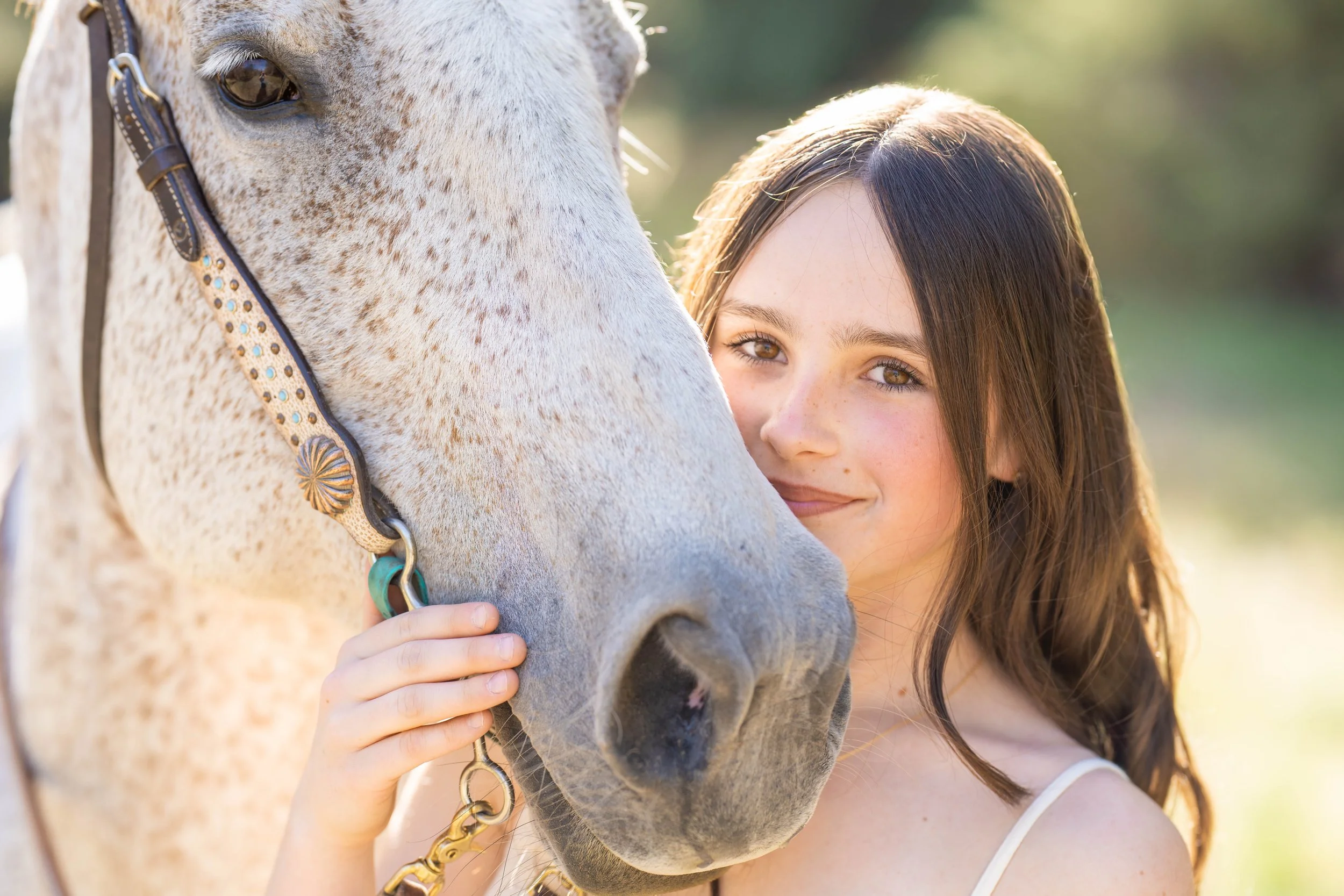 A young woman with brown hair and light skin is standing close to a white horse with light brown spots. She is holding the horse's halter strap with her left hand and smiling softly. The background is blurry, with green foliage.