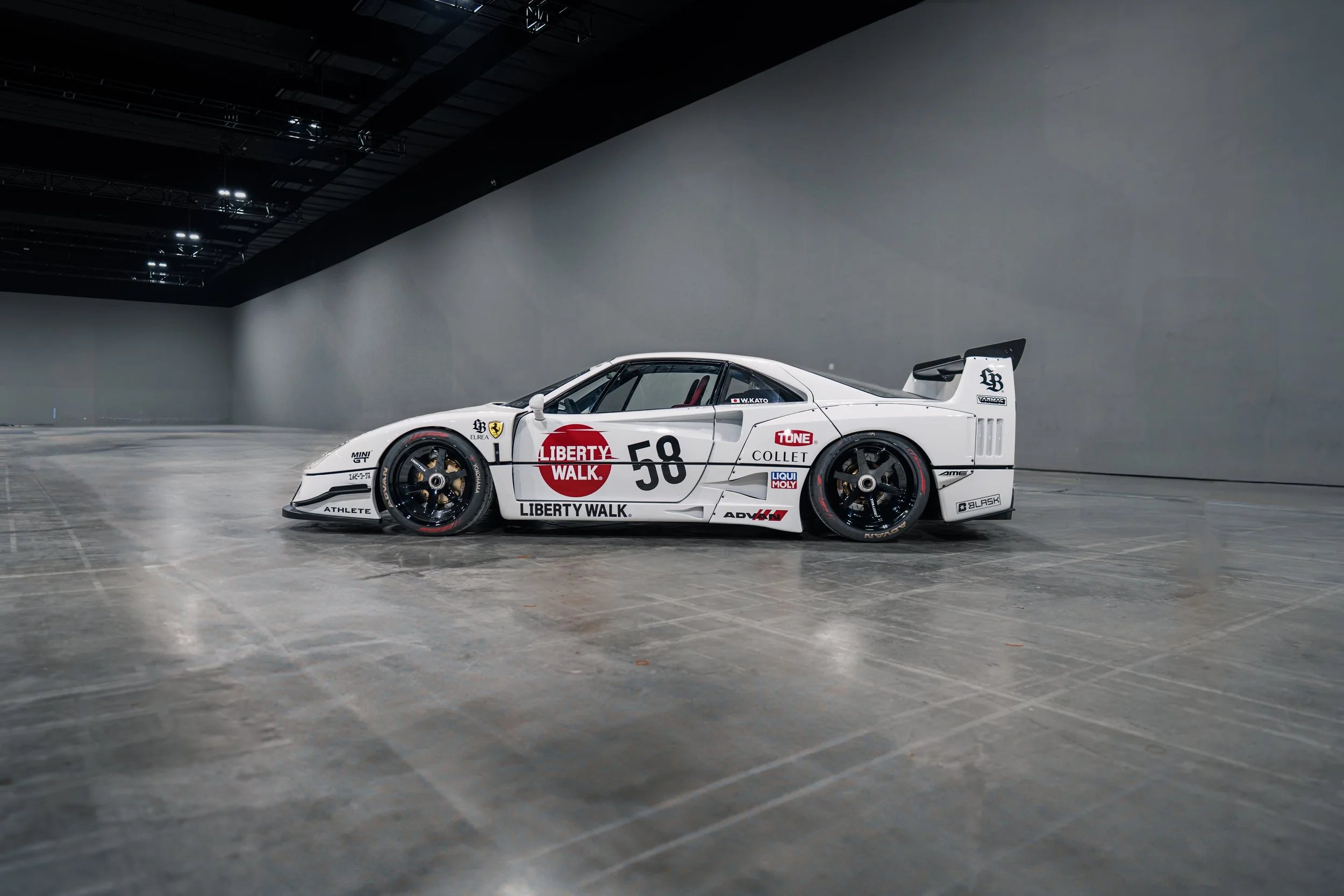 The front of a racing car featuring sponsor logos, including a red Liberty Walk logo and a large white 'CHAMPAGNE COLLET' logo, with the car number 50 visible, partially showing yellow and black caution tape on the floor nearby.
