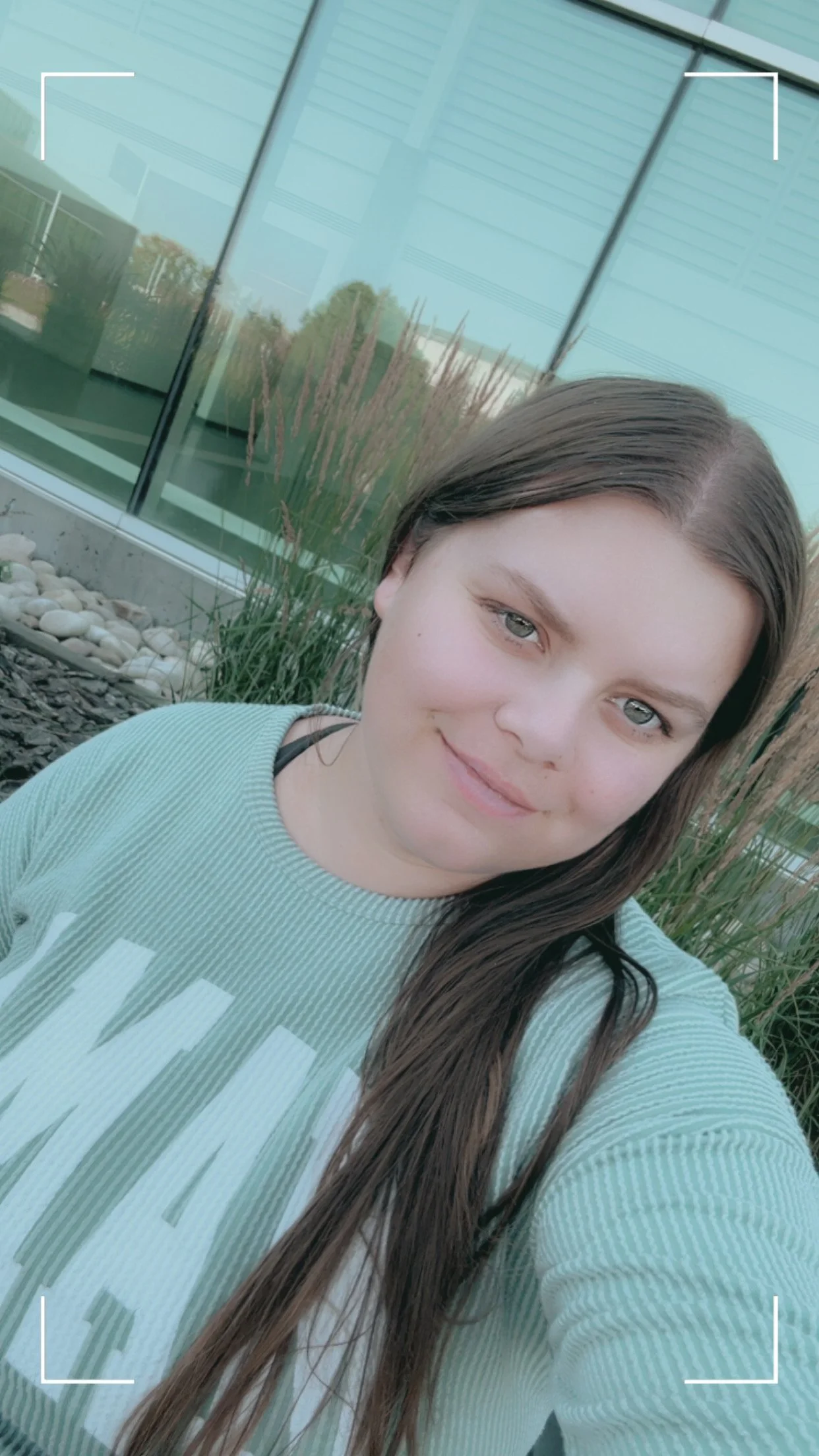 A young woman with long brown hair and blue eyes taking a selfie outdoors in front of a modern glass building with tall grasses and rocks in the landscaping.