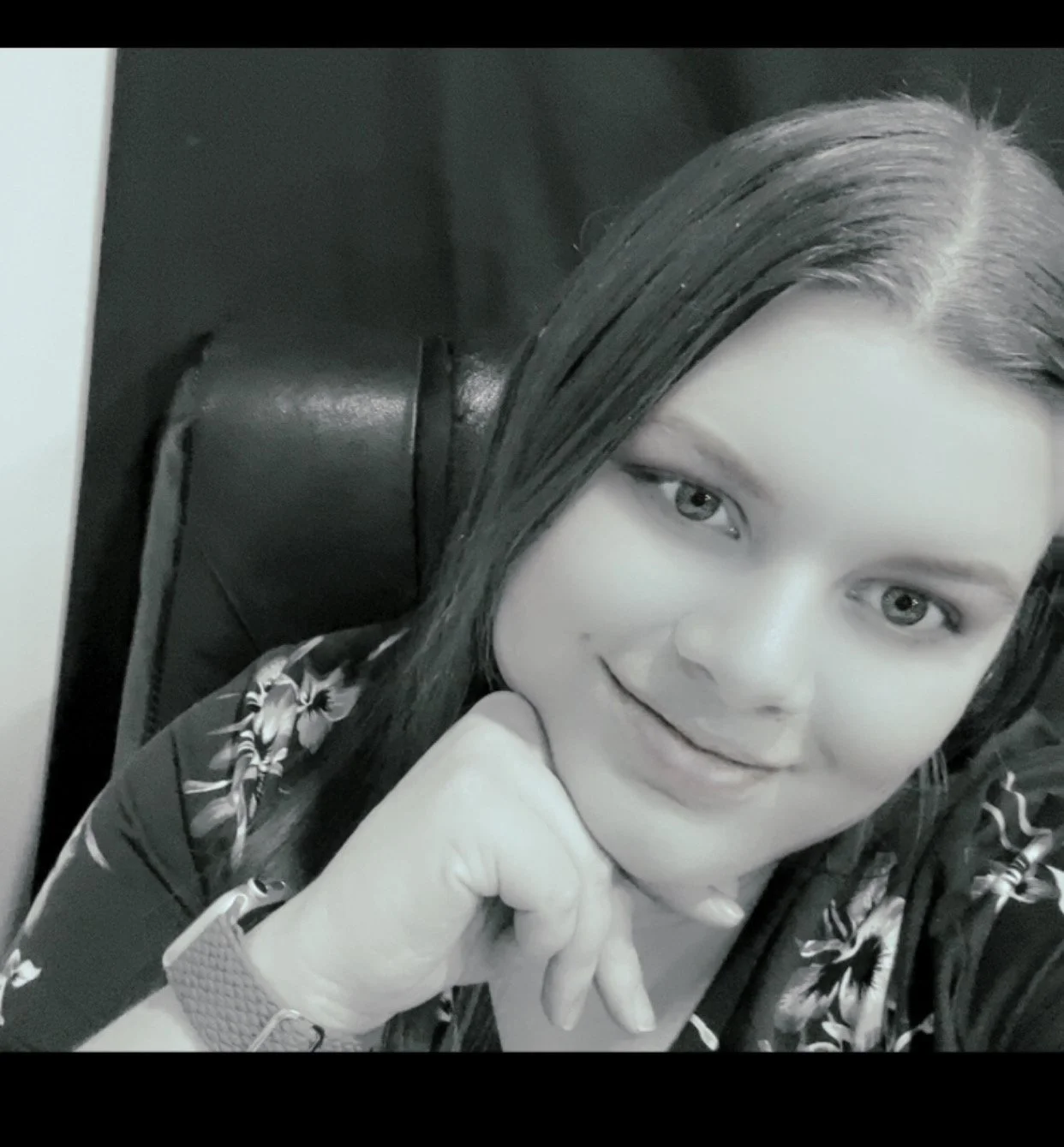 A young woman with long dark hair, resting her chin on her hand, sitting in a black office chair, wearing a floral-patterned top, and smiling slightly at the camera.