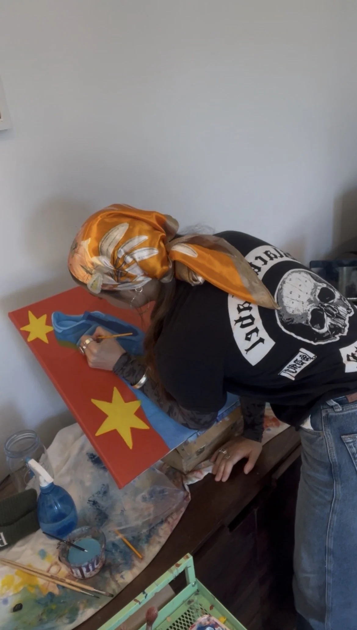 Person with a headscarf painting a flag of China on a canvas, wearing a black T-shirt with a skull design, standing at a table with painting supplies.