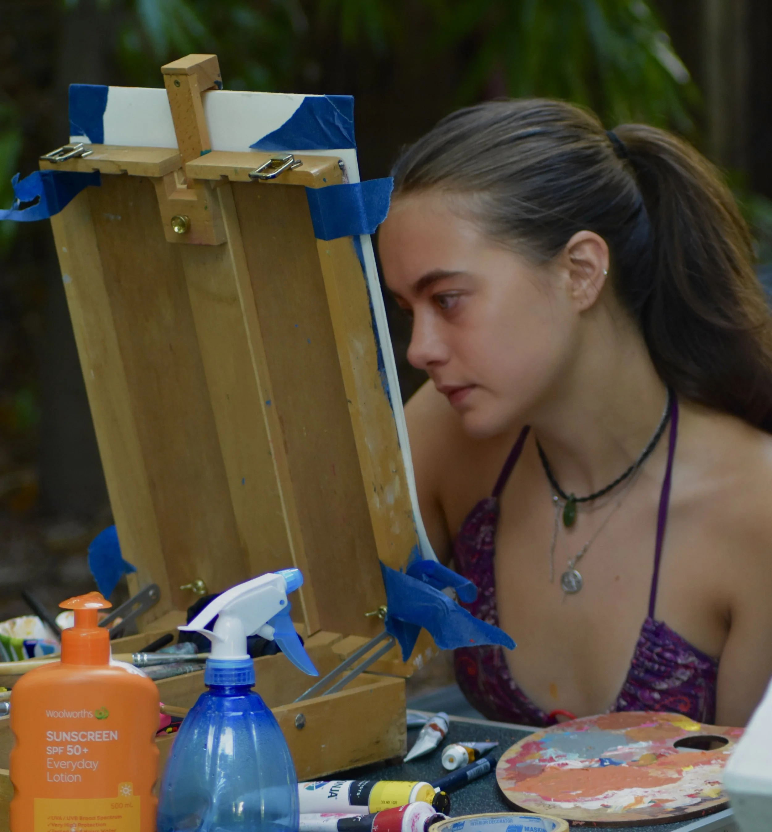 A young woman with long brown hair tied back, looking intently at a painter's palette with paint brushes and tubes around her, during an outdoor painting session.