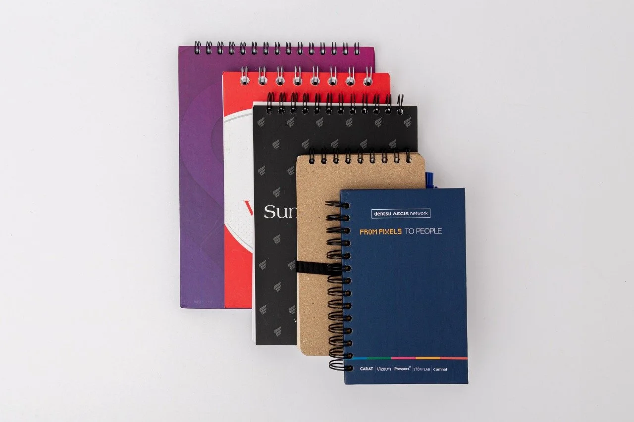 A stack of five spiral notebooks on a white surface, with varying colors and sizes, including purple, red, black, brown, and blue covers.