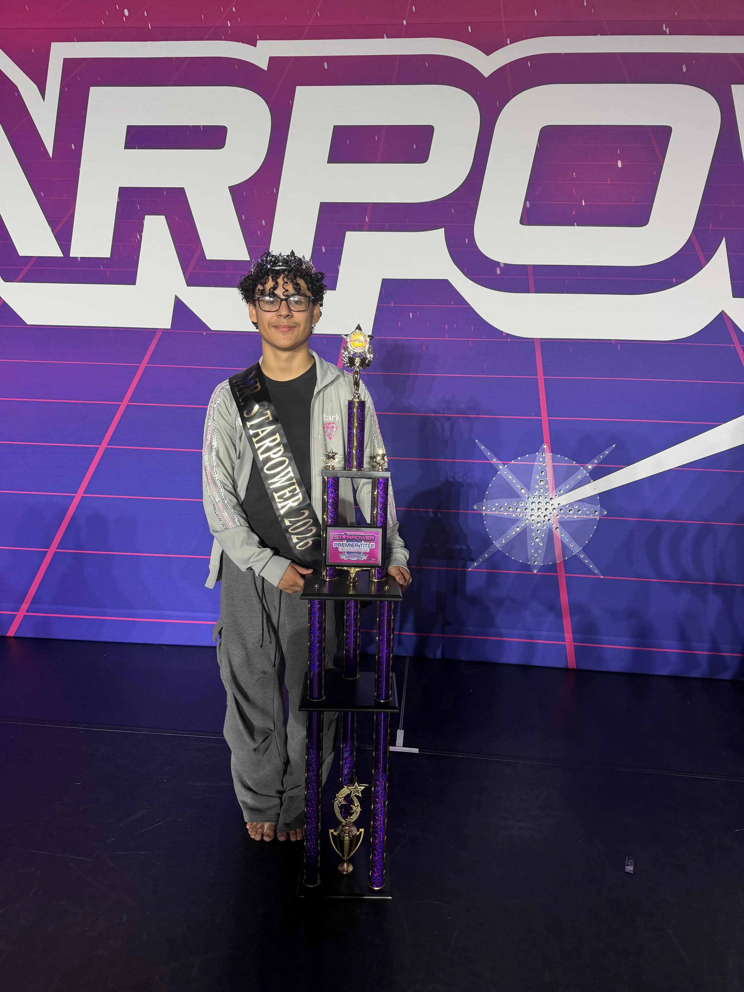 A young person standing next to a tall trophy with a large star on top, in front of a purple, pink, and black backdrop with a star graphic and large text. The person is wearing glasses, a crown, a silver and black jacket, a sash that says 'Starpower 2020', and gray pants.