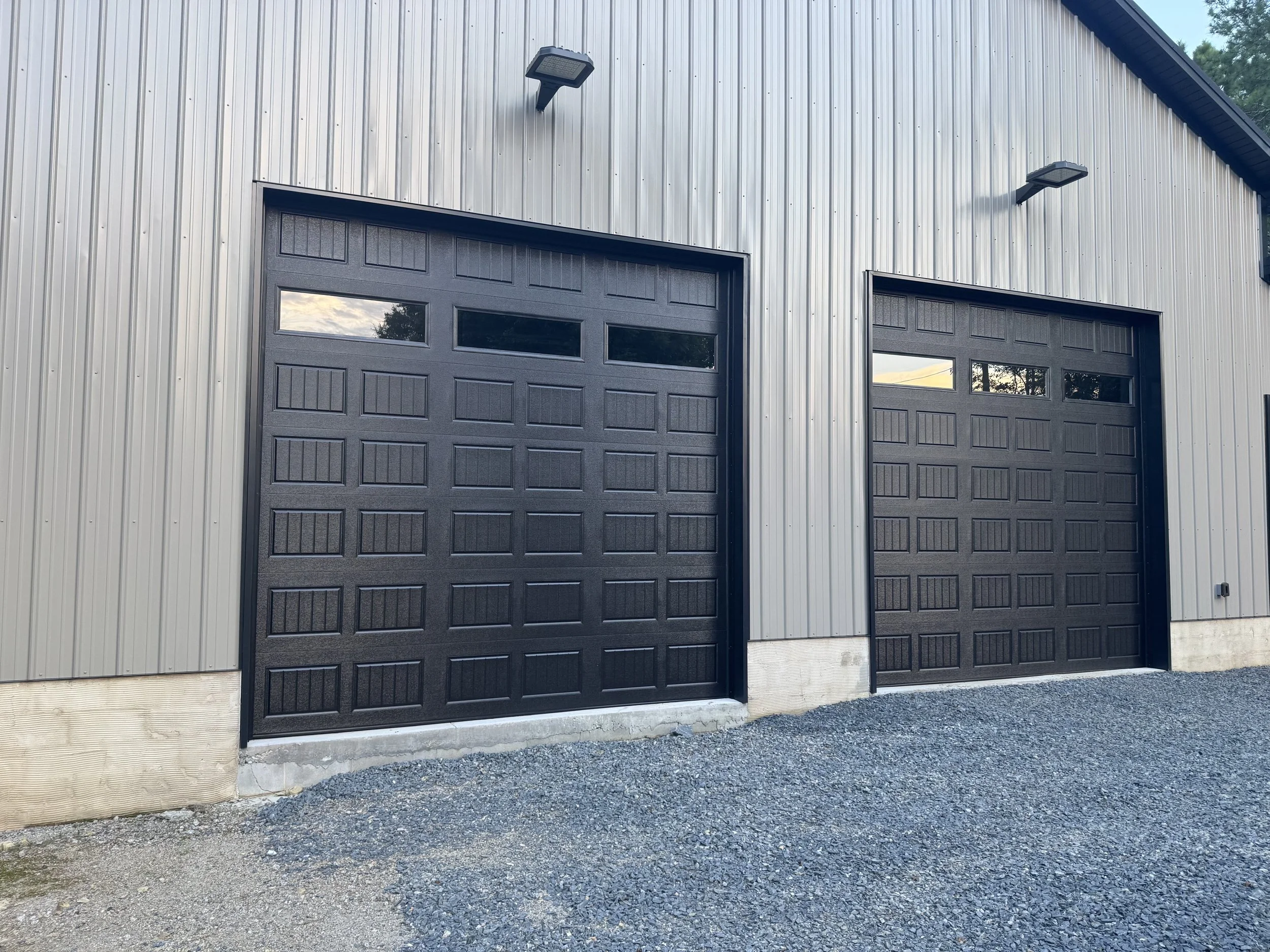 Two black garage doors with windows at the top, mounted on a metallic building with outdoor lights above each door, and gravel on the ground in front.