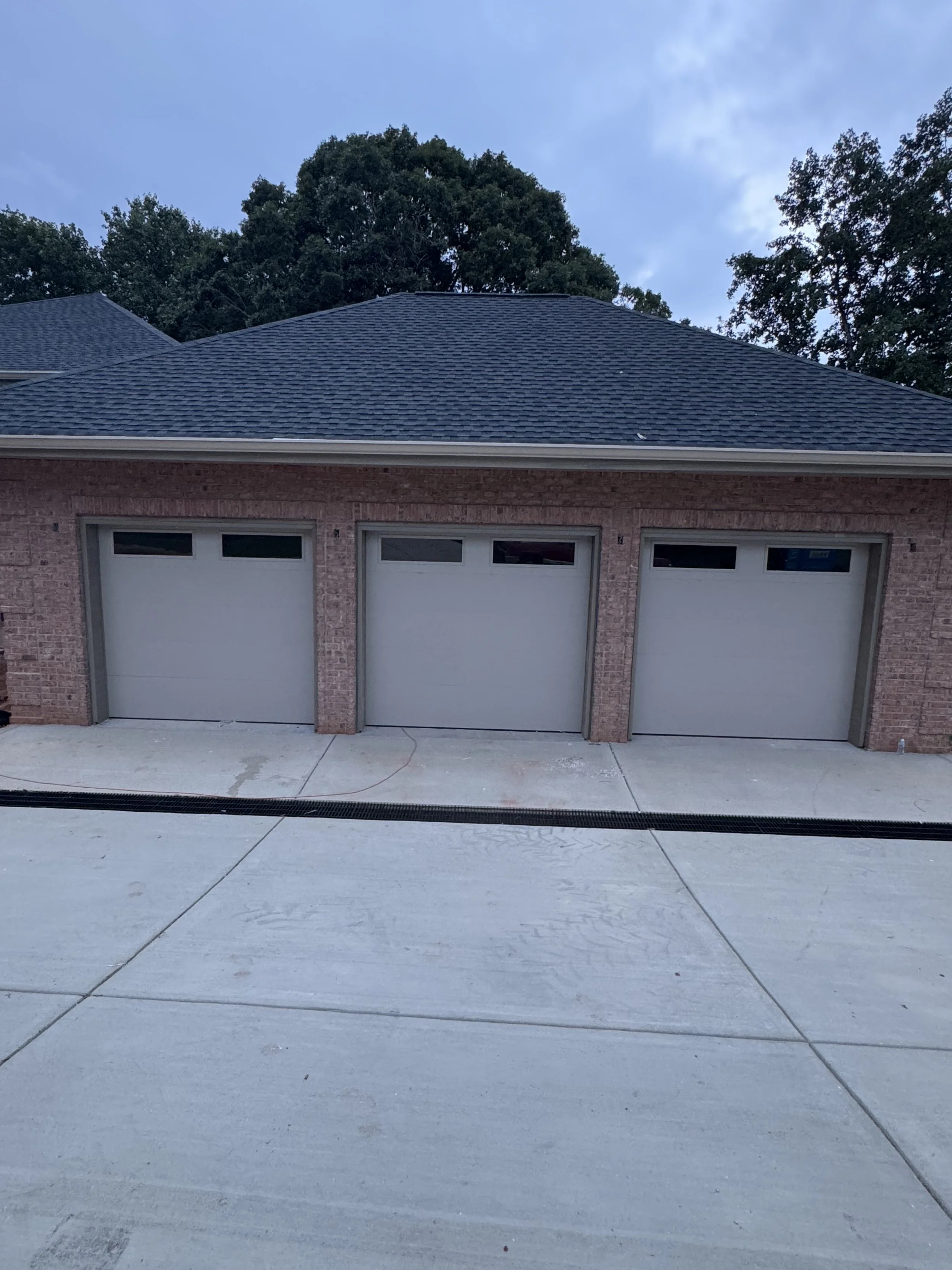 Three closed garage doors on a brick house with a shingled roof, concrete driveway, and trees in the background