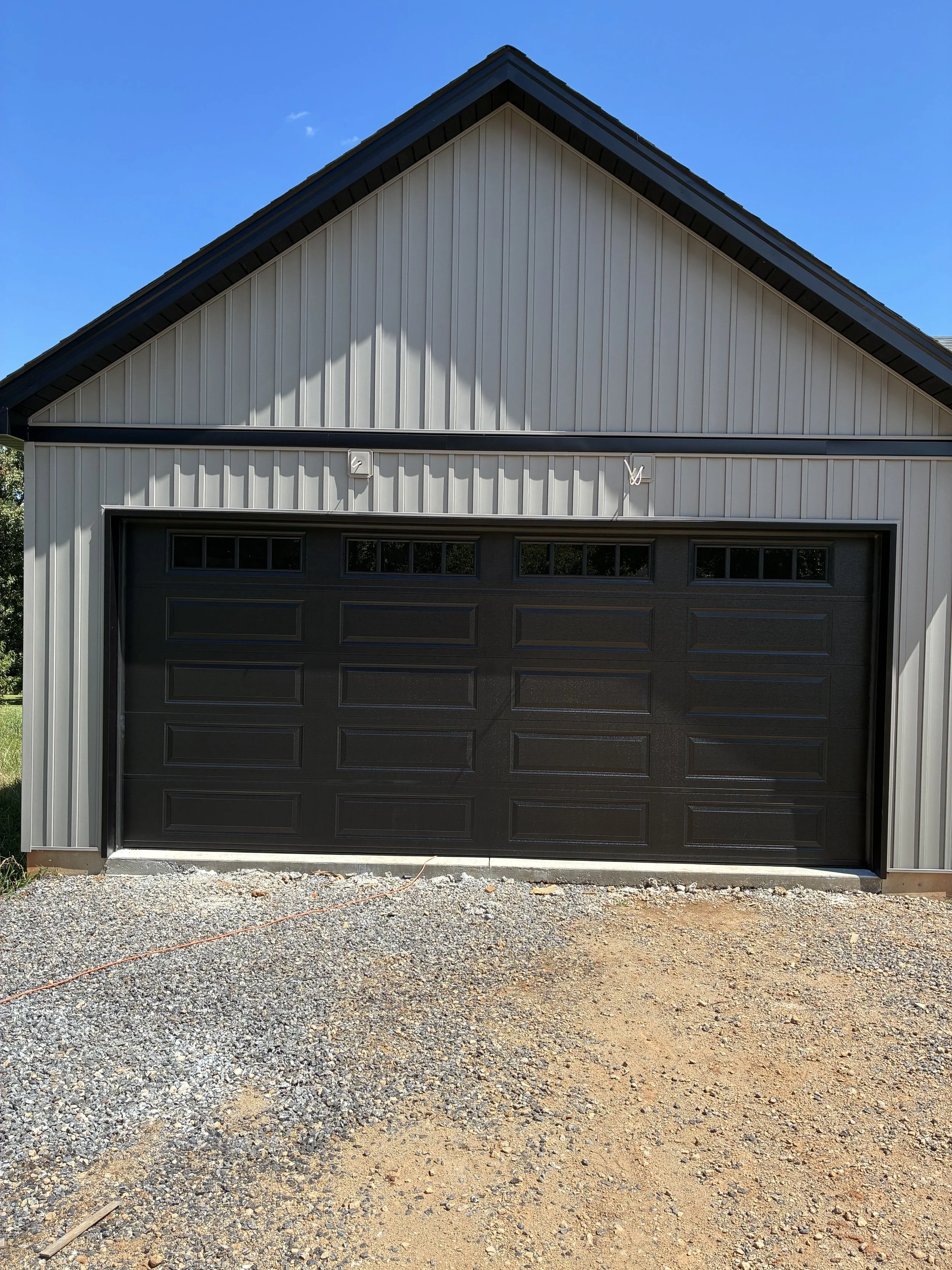 Front view of a metal garage with a black roll-up door and small windows at the top, under a metal-paneled roof, on a gravel driveway.