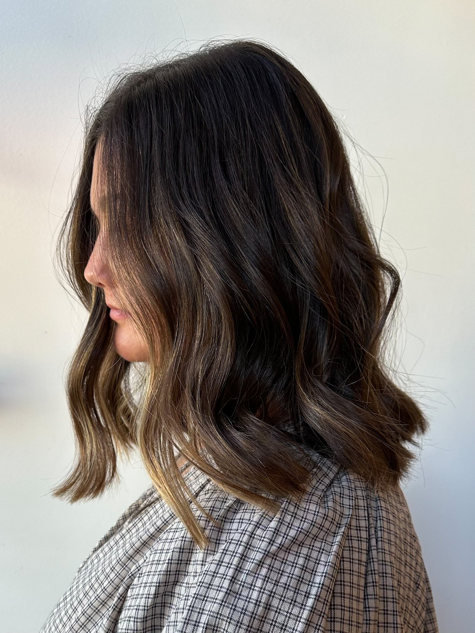 Side view of a woman with shoulder-length wavy brown hair with subtle highlights, wearing a checkered shirt against a plain background.