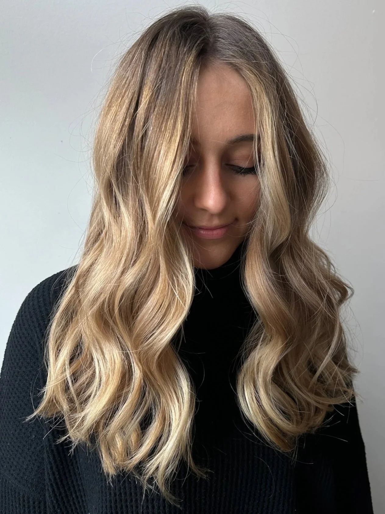 A woman with long, wavy blonde hair, wearing a black top, looking down with her eyes closed.