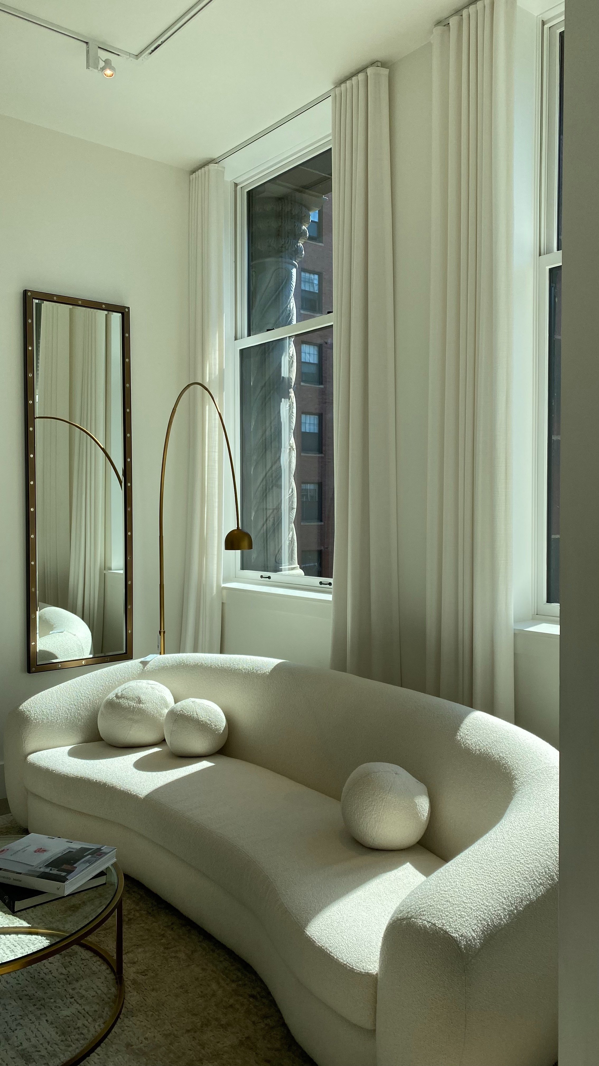 Bright living room with a cream curved sofa, a tall gold arc floor lamp, two large windows with white curtains, a rectangular mirror reflecting part of the room, and a small round coffee table with magazines.