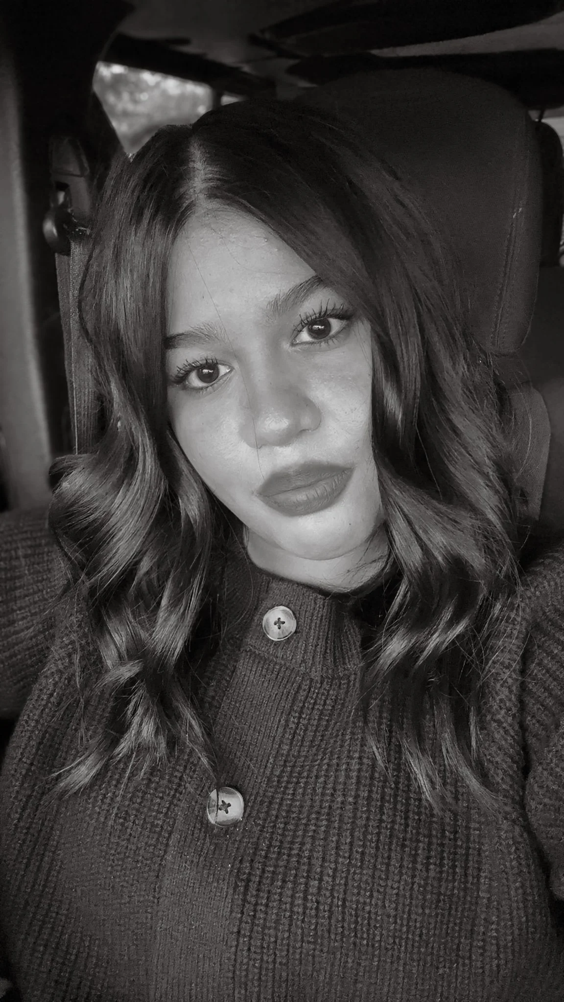 Black and white photograph of a young woman with wavy hair, seated in a car, wearing a button-up sweater.