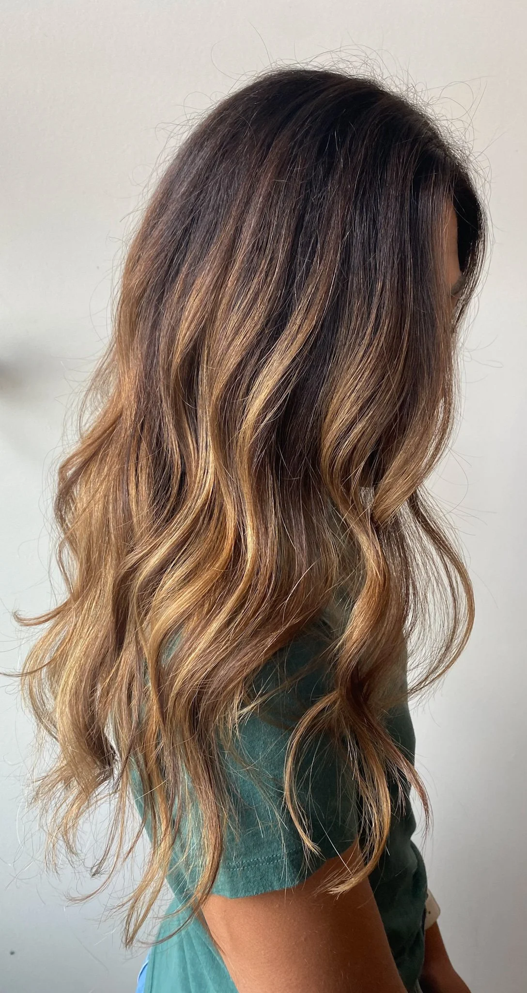 Side view of a woman with long, wavy hair with balayage coloring in shades of dark brown, light brown, and blonde, wearing a teal shirt.