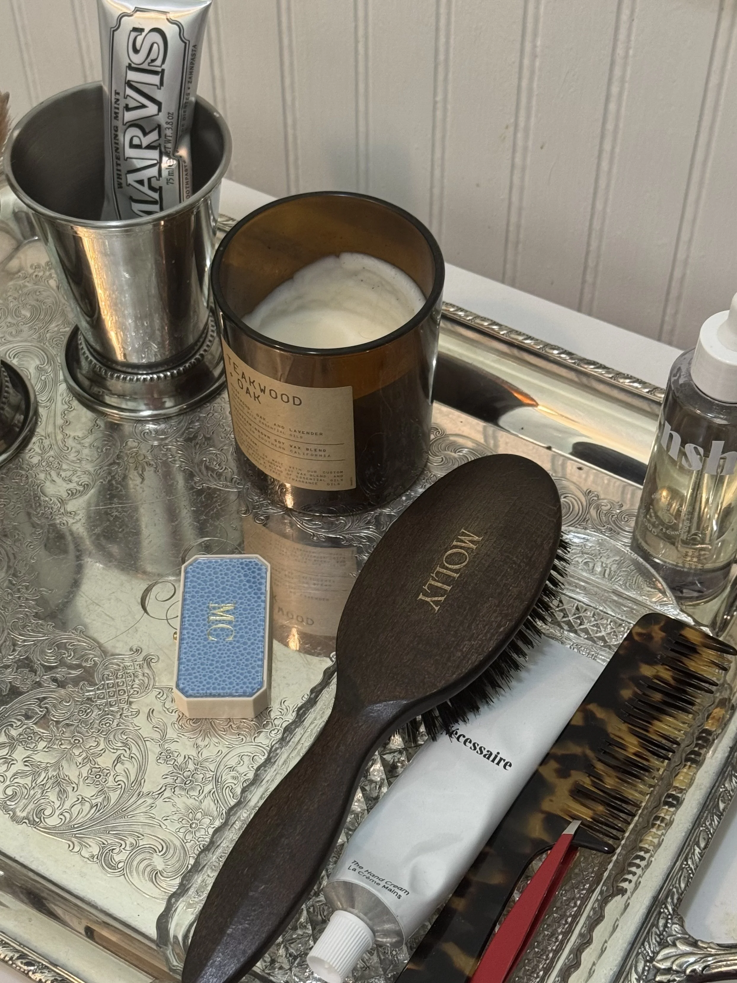 A silver tray holding grooming and personal care items, including a wooden hairbrush with 'MOLLY' engraved, a tortoiseshell hair comb, hand sanitizer, a tube of hand cream, a small blue and white box, a container of candles, an ice mint candy bar, and a silver cup.