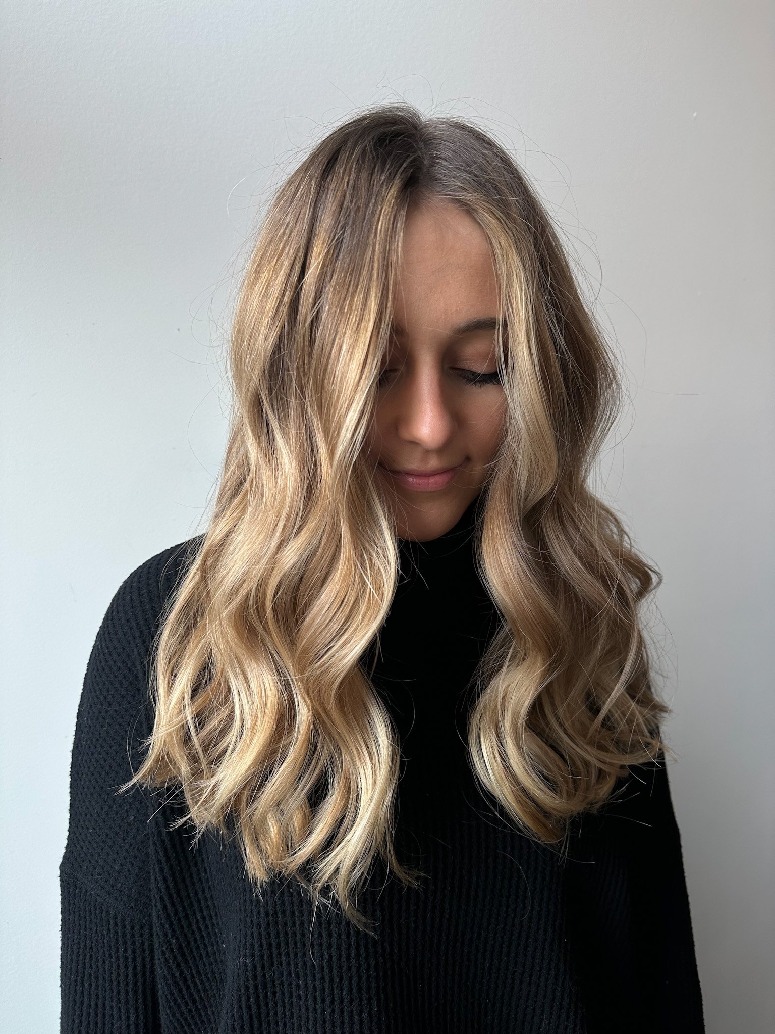 A woman with long, wavy blonde hair looking downward with her eyes closed, wearing a black textured top, standing against a plain light gray background.