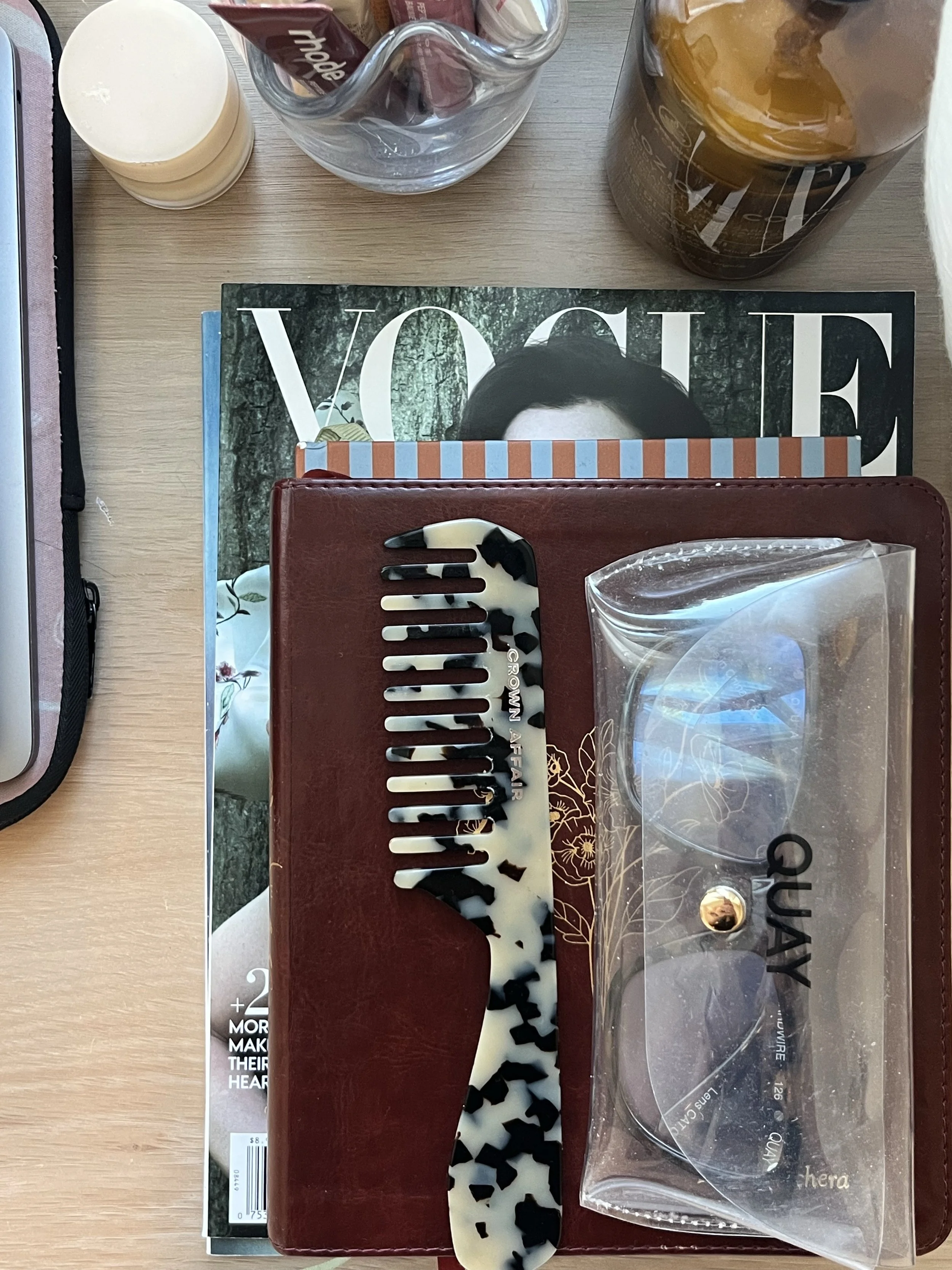 Top view of a desk with a Vogue magazine, a red wallet, a tortoiseshell hair comb, a pair of sunglasses in a clear case, a container of honey, a glass jar with tea bags, and a white cosmetic container.