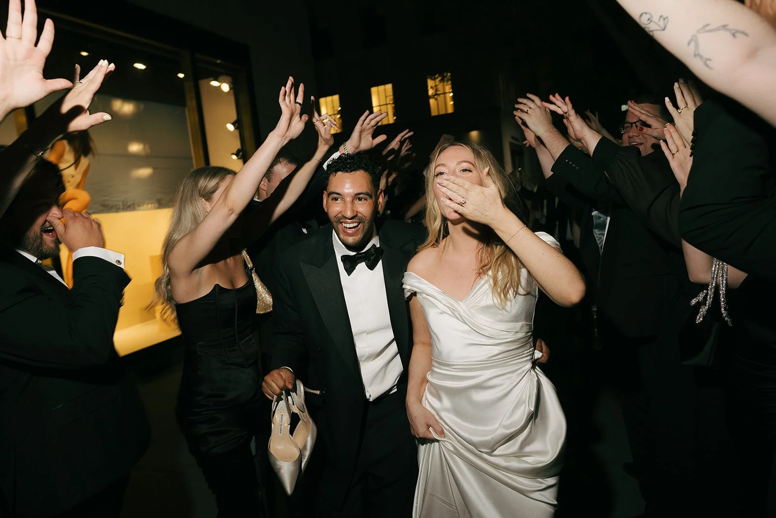 A wedding celebration where a bride and groom walk through a tunnel of smiling guests, with guests raising their hands and cheering.