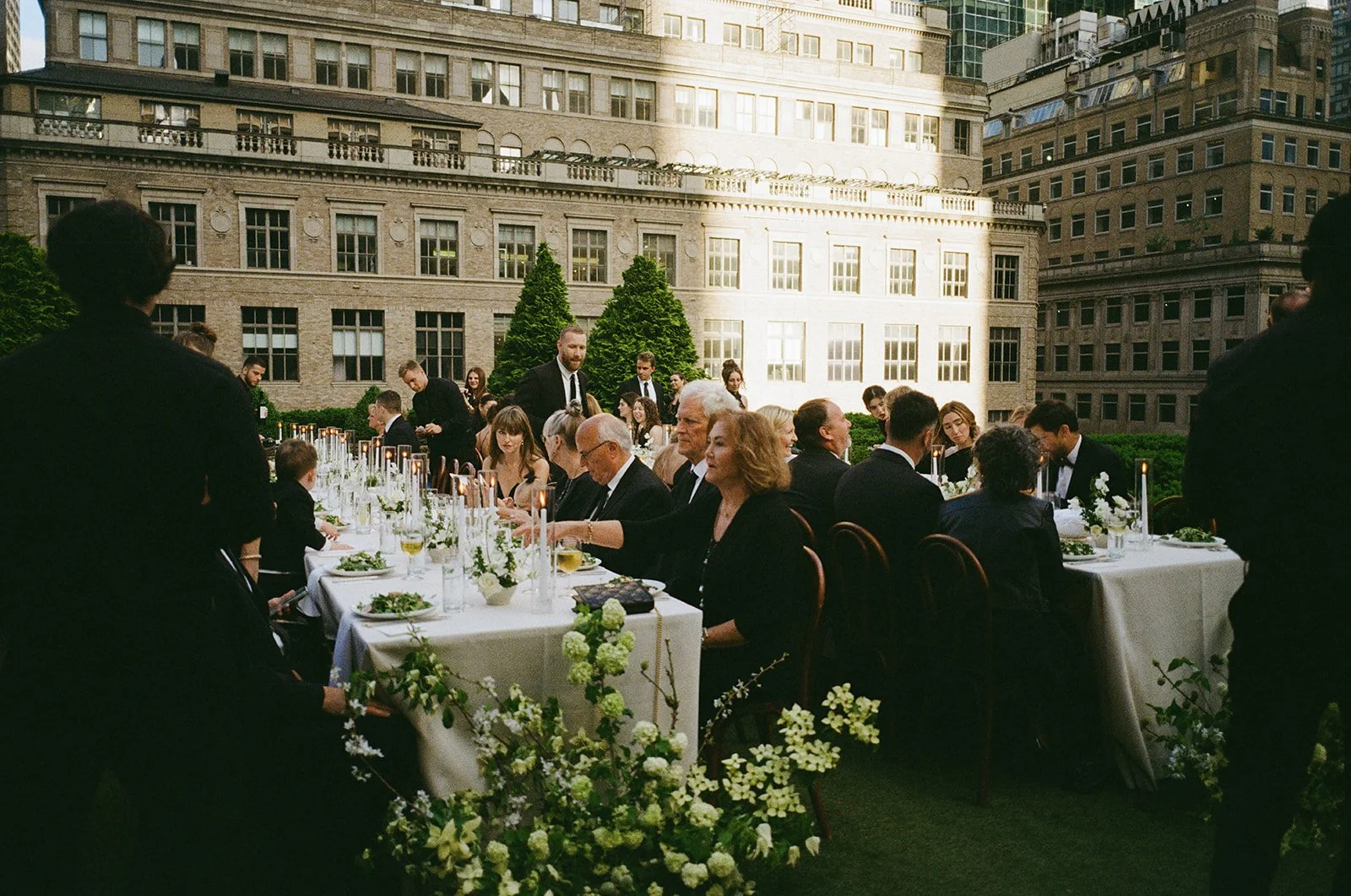 A large outdoor dinner event on a rooftop with people dressed in formal attire, seated along a long banquet table decorated with flowers and candles, against a backdrop of tall building facades and greenery.