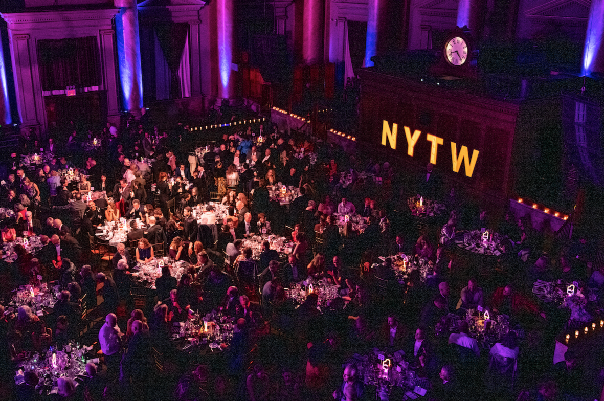 A large crowd of people seated at round tables, attending an event in a grand hall with purple lighting. There is a large sign with the letters "NYTW" illuminated on the wall.