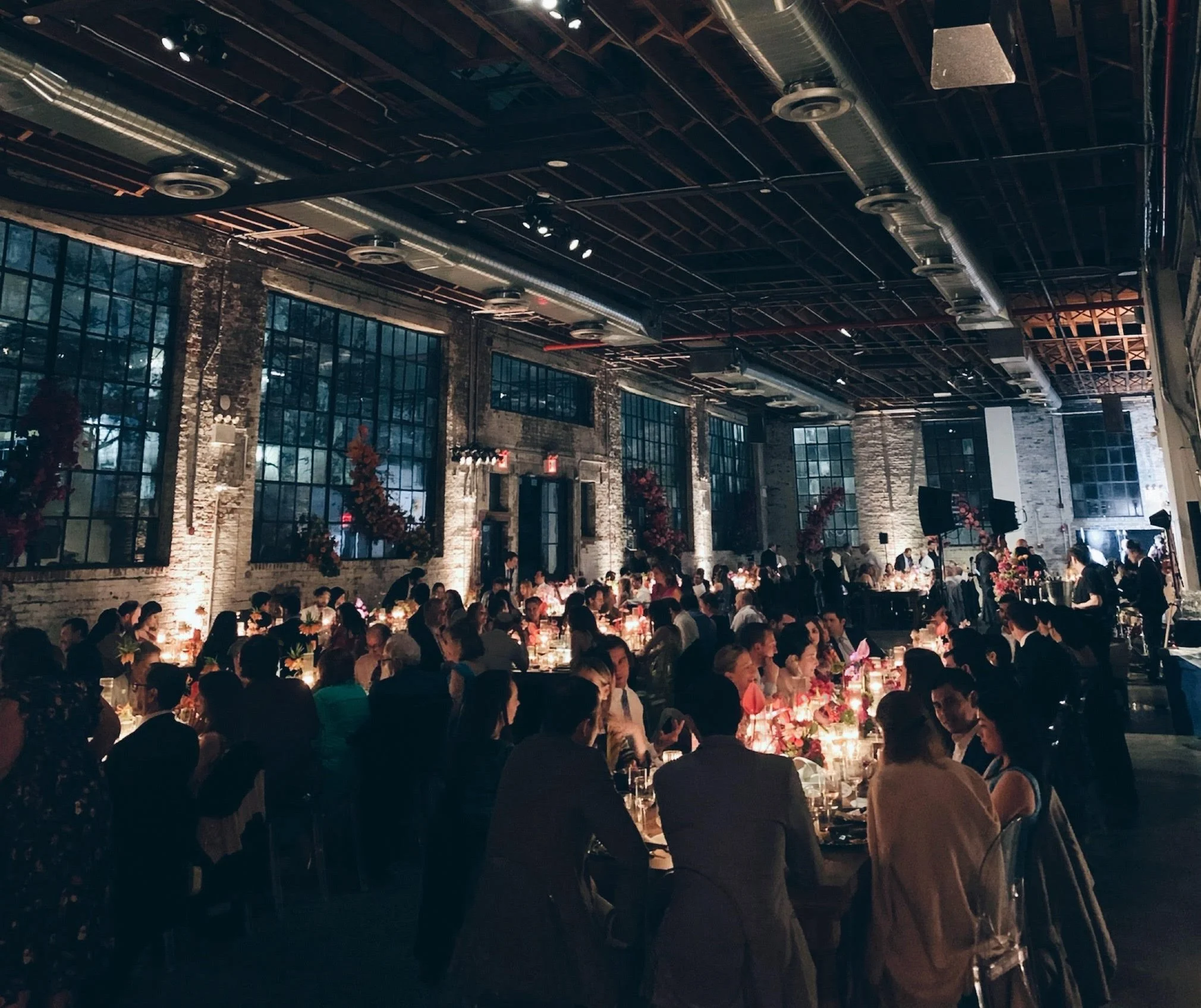 A crowded indoor event with people seated at long tables decorated with flowers and candles, in a venue with exposed brick walls and large industrial-style windows.