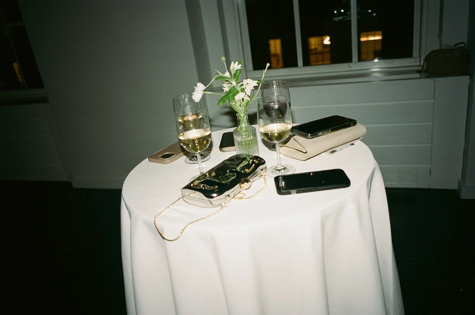 A round table with a white tablecloth, three wine glasses filled with white wine, a small bouquet of white flowers in a glass vase, two smartphones, a beige clutch, a black clutch, and a beige napkin. The background shows a window with night view.