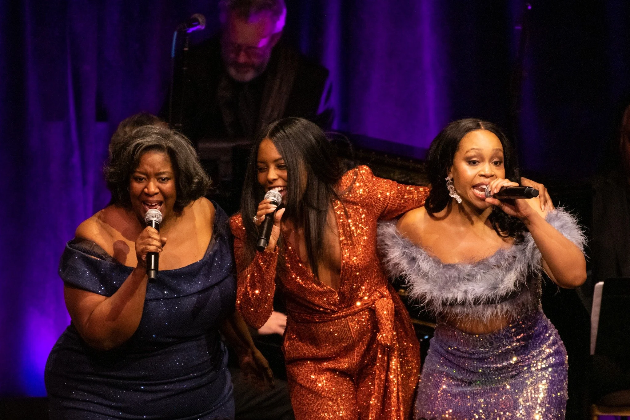 Three women are singing into microphones on stage at a lively event, with a pianist in the background.