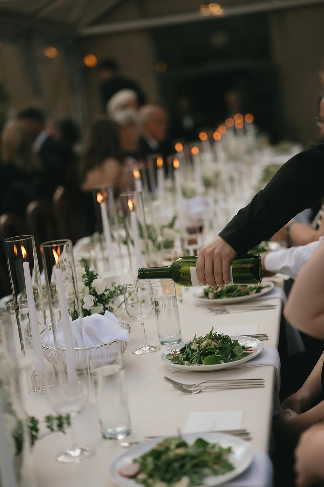 A person pouring wine into a glass at a formal dinner party with a long table set with plates, silverware, candles, and floral arrangements.