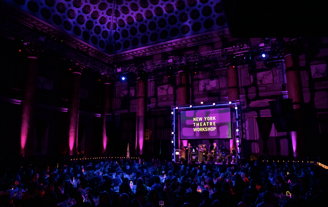 An auditorium hosting the New York Theatre Workshop with stage lights and a band performing. The audience is seated at tables.