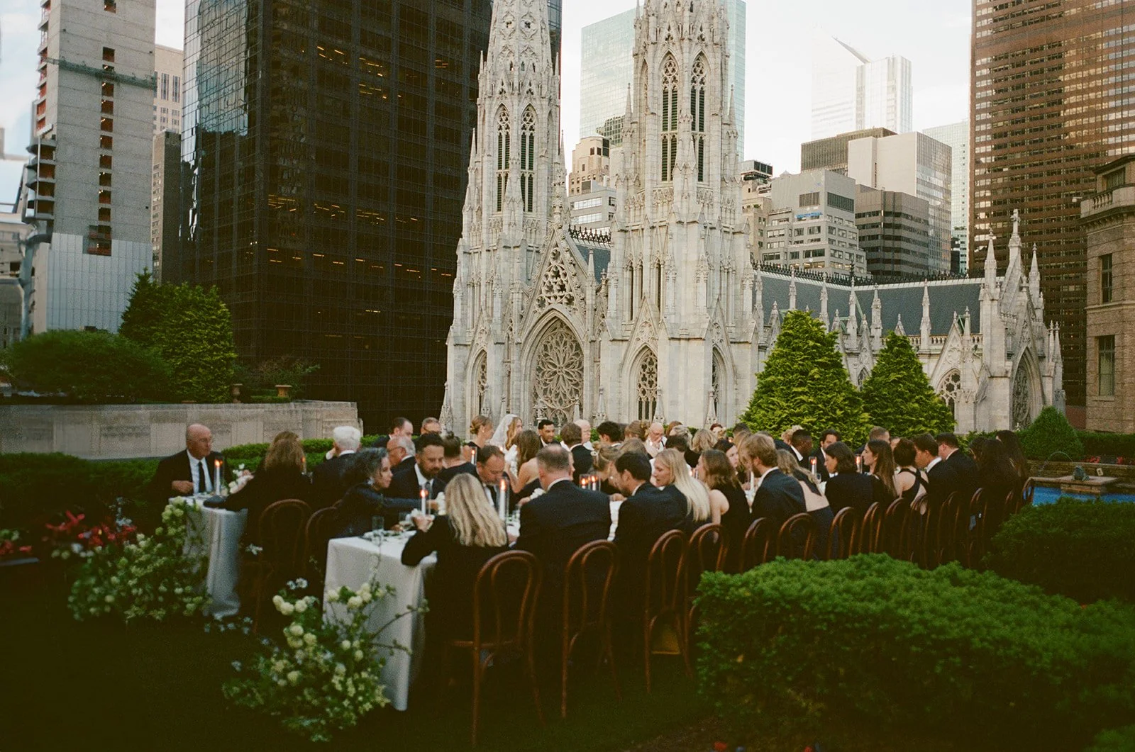 Outdoor evening wedding reception on a rooftop patio with a gothic-style church in the background, guests seated at long tables with white tablecloths and floral centerpieces.