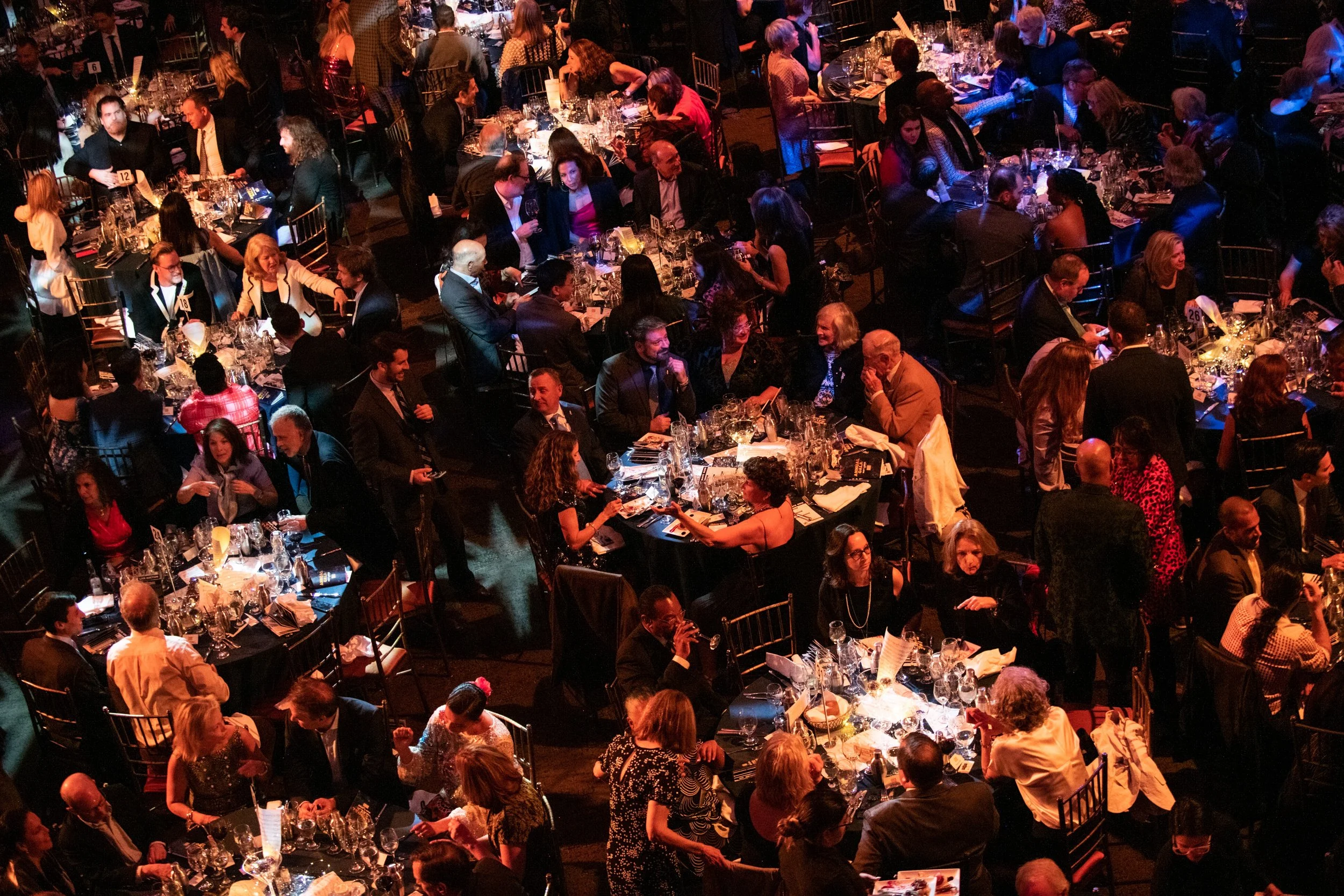 An overhead view of a formal dinner event with many round tables filled with people dressed in formal attire, engaged in conversations and enjoying food and drinks in a dimly lit setting.