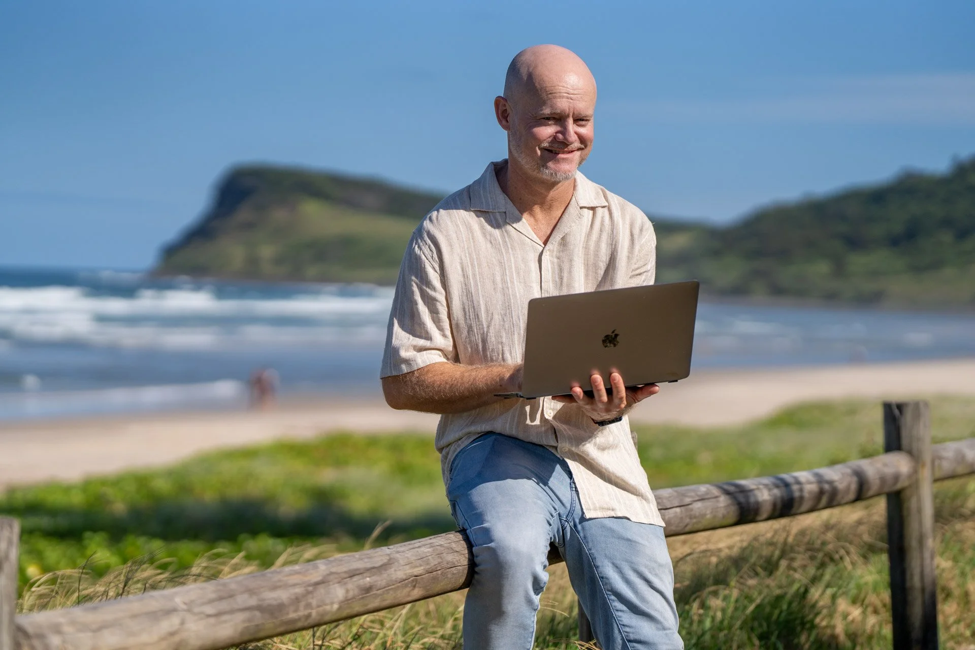 A man with a bald head and a goatee, wearing a beige striped shirt and jeans, is sitting on a wooden fence at the beach, holding a laptop and smiling. The background features the ocean, sandy beach, and a green hill under a clear blue sky.