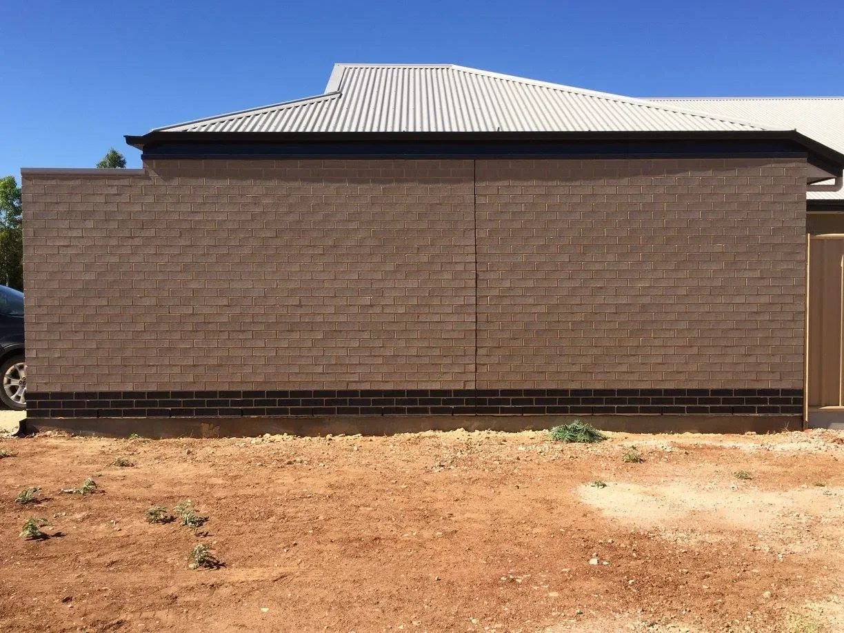Brown brick wall with a metal roof, part of a building, on a patch of dirt ground.