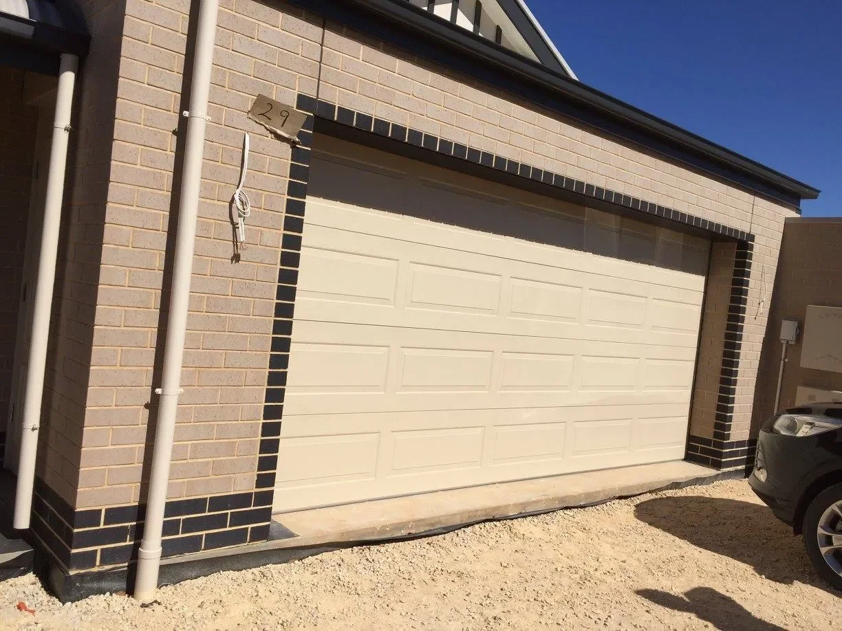 A closed white garage door attached to a brick house with black and beige bricks. A small sign with the number 29 is above the garage door. To the right, a black car is partially visible. The ground is covered with gravel, and the sky is clear and bl