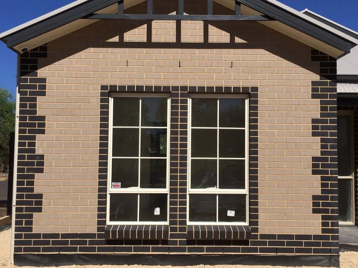 Front view of a brick house with two large windows, light brown and black bricks, and a gable roof.