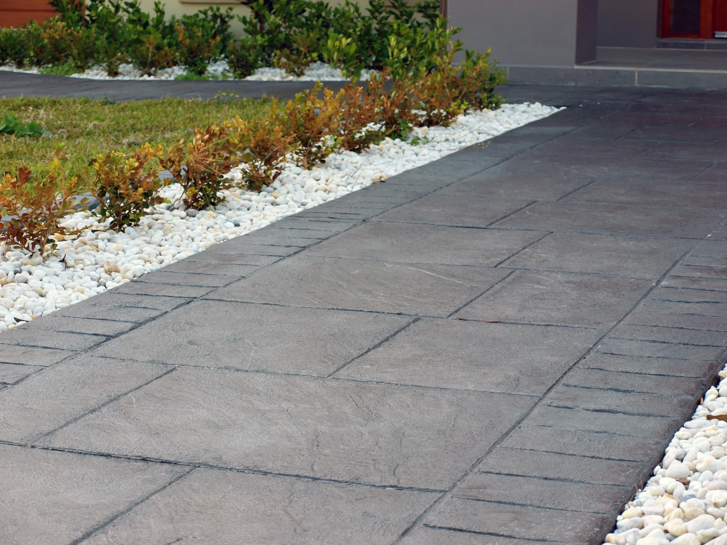 Close-up view of a paved footpath with decorative bedding of small white stones and low green and brown bushes in the background.