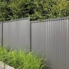 A metal privacy fence with vertical panels, situated next to green grass and bushes, with trees in the background.