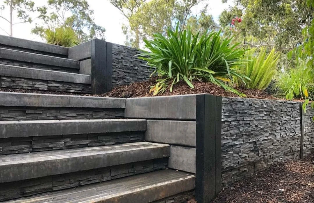 Outdoor staircase with black stone walls and wooden steps, bordered by landscaped plants and trees.
