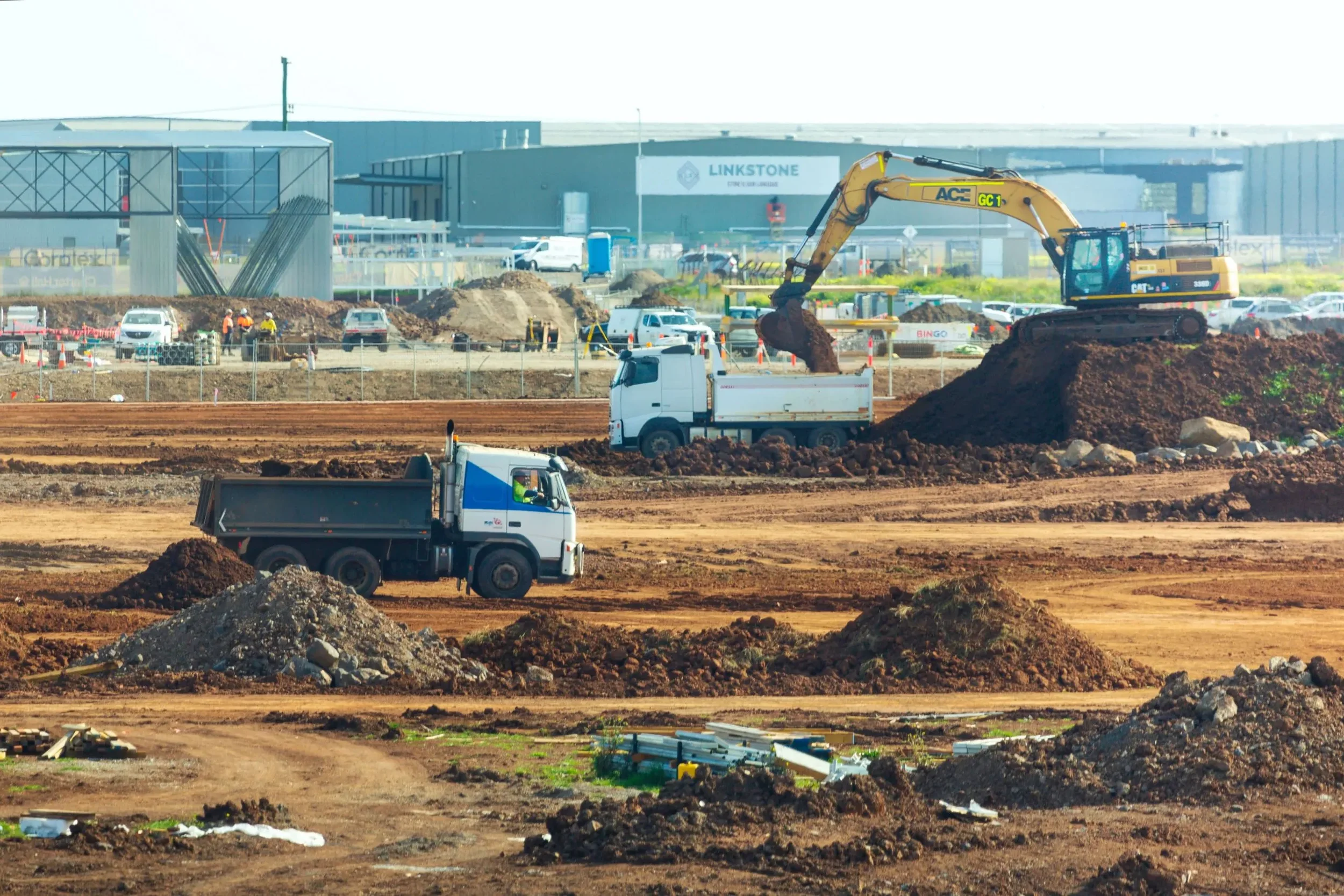 Construction site with trucks and excavators working on the dirt, with industrial buildings and parked cars in the background.