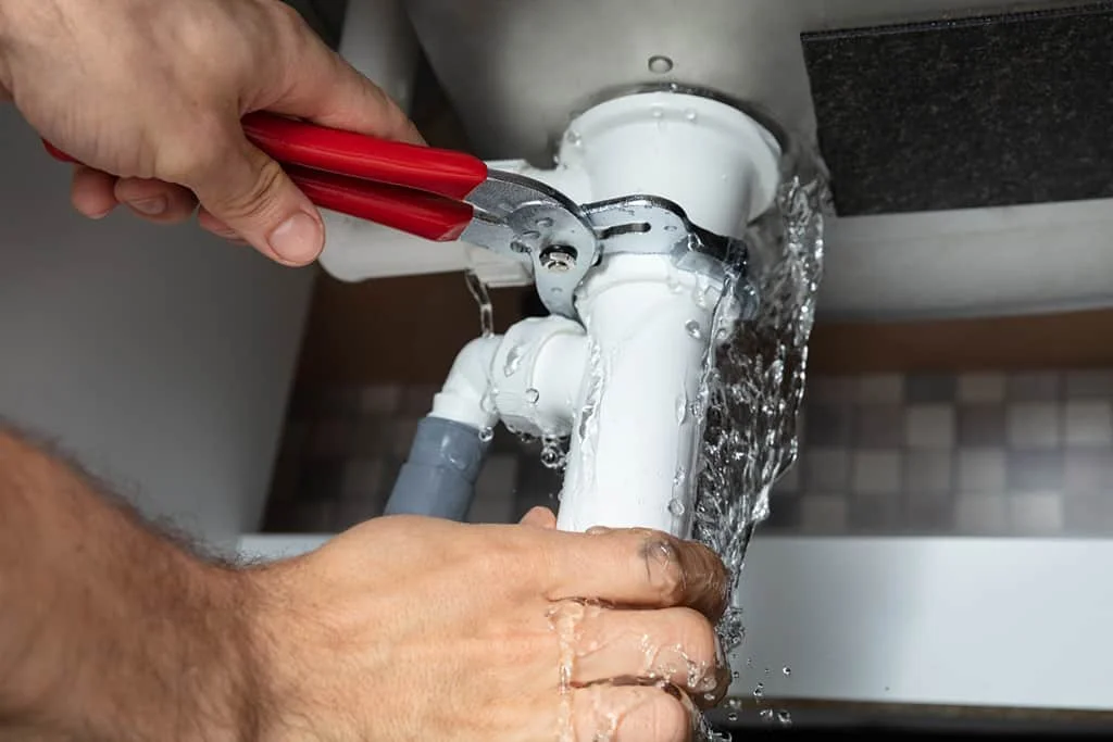 Hands tightening the seal around a white PVC pipe as water leaks from the sink above.