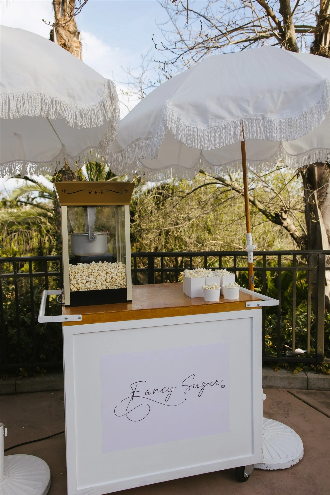 A popcorn stand called 'Fancy Sugar' with white umbrellas providing shade, set outdoors with trees in the background.