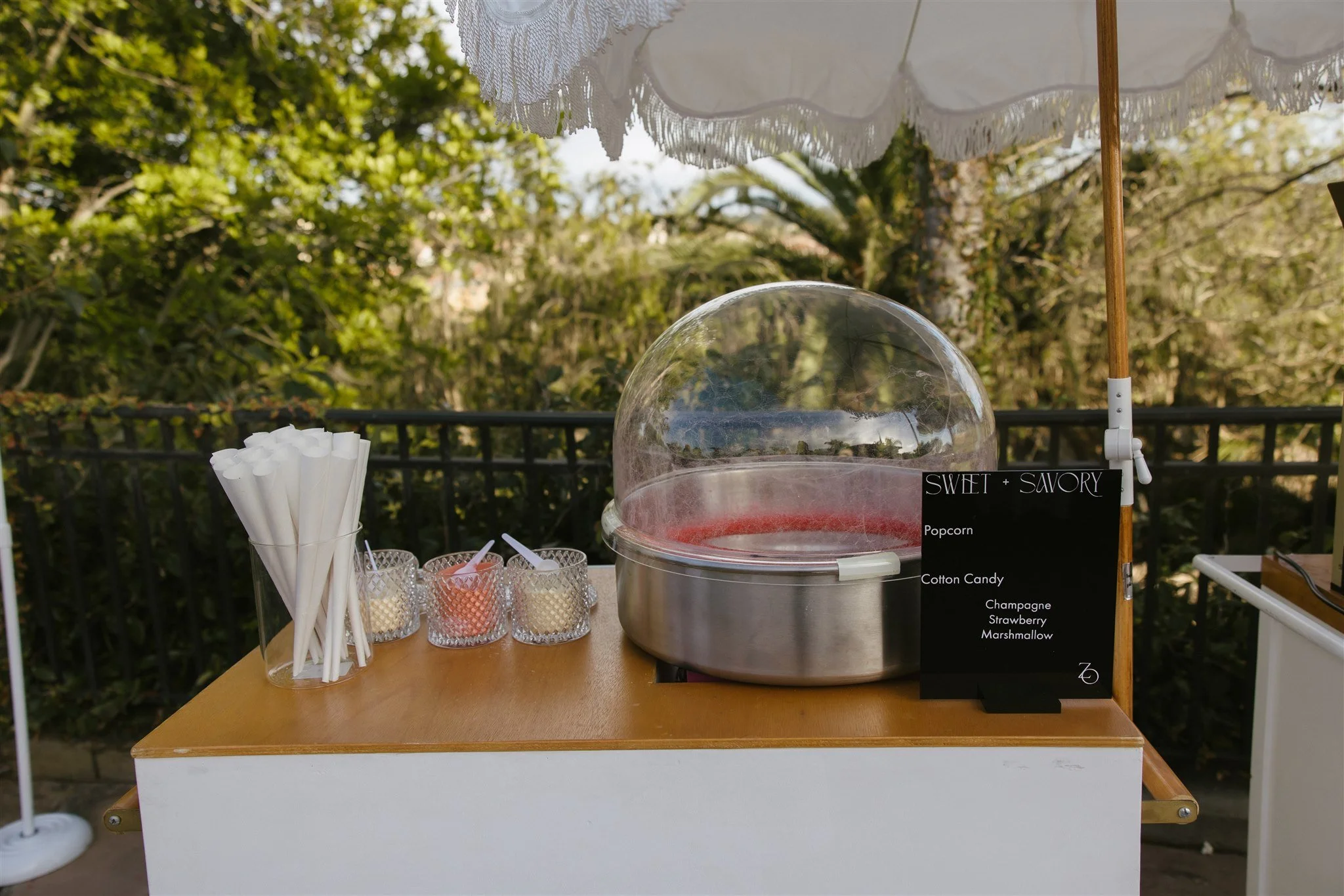 A cotton candy and popcorn stand outdoors with trees and a black fence in the background. The stand features a cotton candy machine with pink cotton candy inside, a black sign listing flavors including popcorn, cotton candy, champagne, strawberry, an